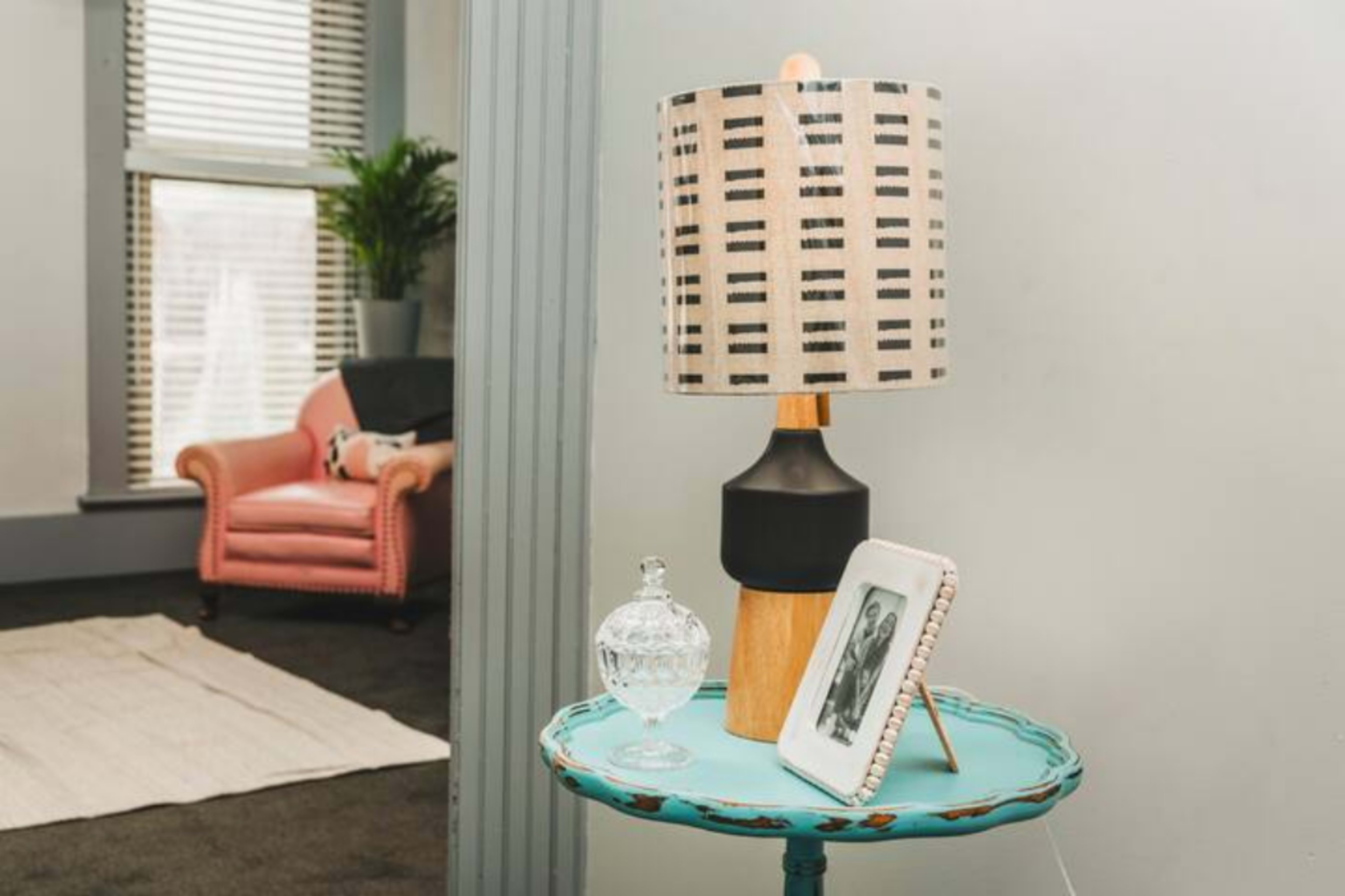 The image features a stylish corner of a room with a pink armchair, a small round table holding a lamp, a decorative glass container, and a framed photo.