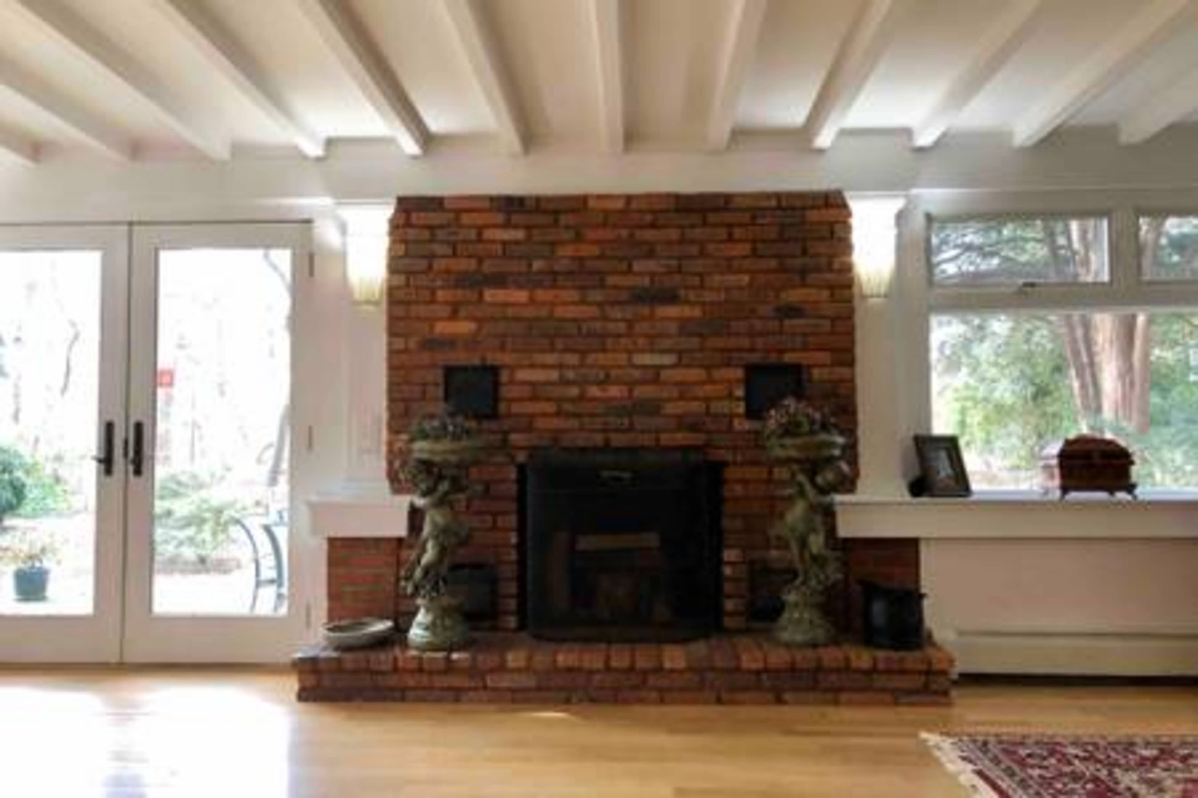 The image shows a brick fireplace with two decorative statues on either side, framed by large windows that provide a view of a wooded area outside.