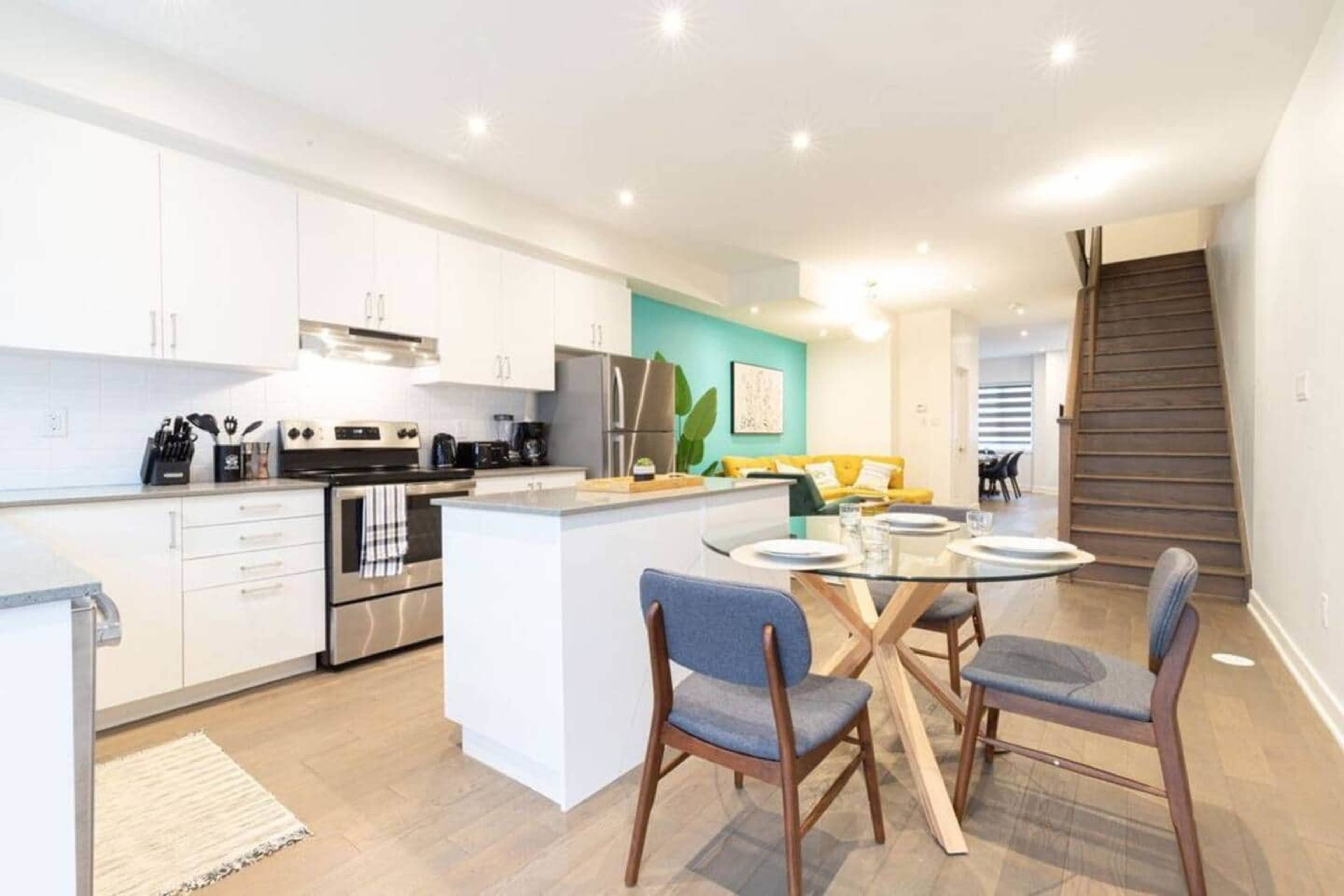 The image shows a modern kitchen and dining area featuring a round table with four chairs, stainless steel appliances, and a staircase leading to an upper level.