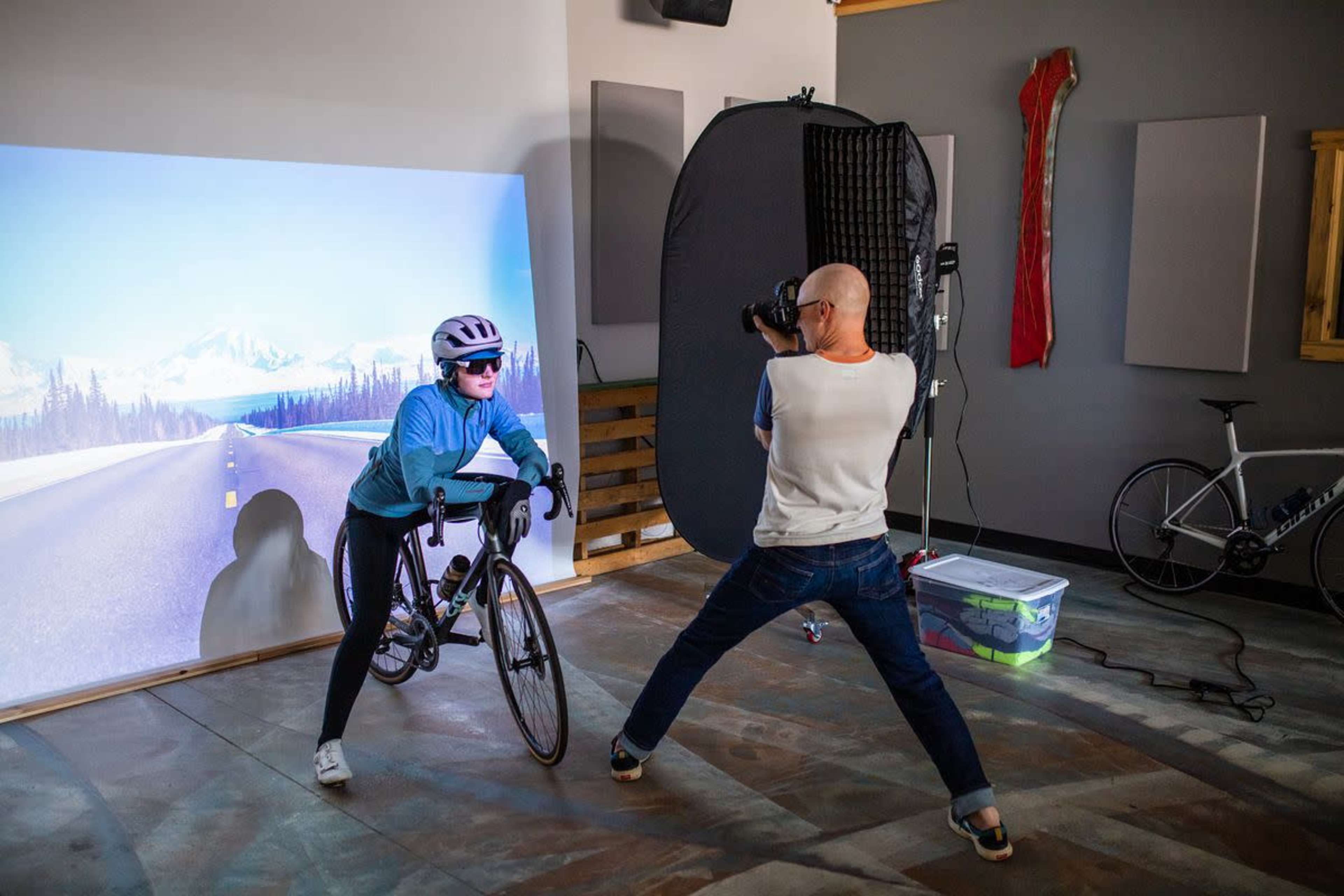 A cyclist poses on a stationary bike in a studio setting while a photographer captures the shot against a backdrop of a scenic road.