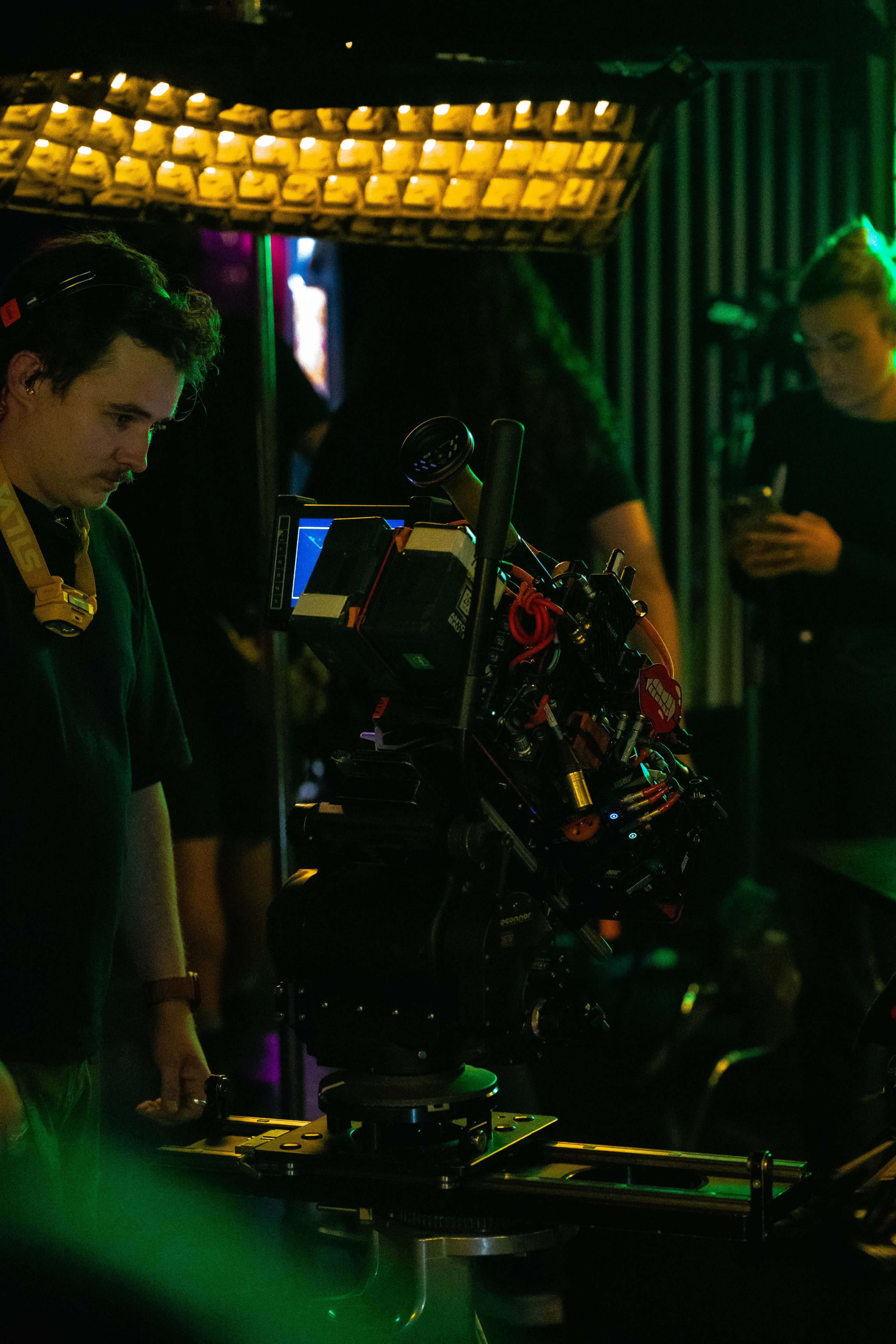 A camera operator adjusts a professional film camera on a tracking rig in a dimly lit studio.