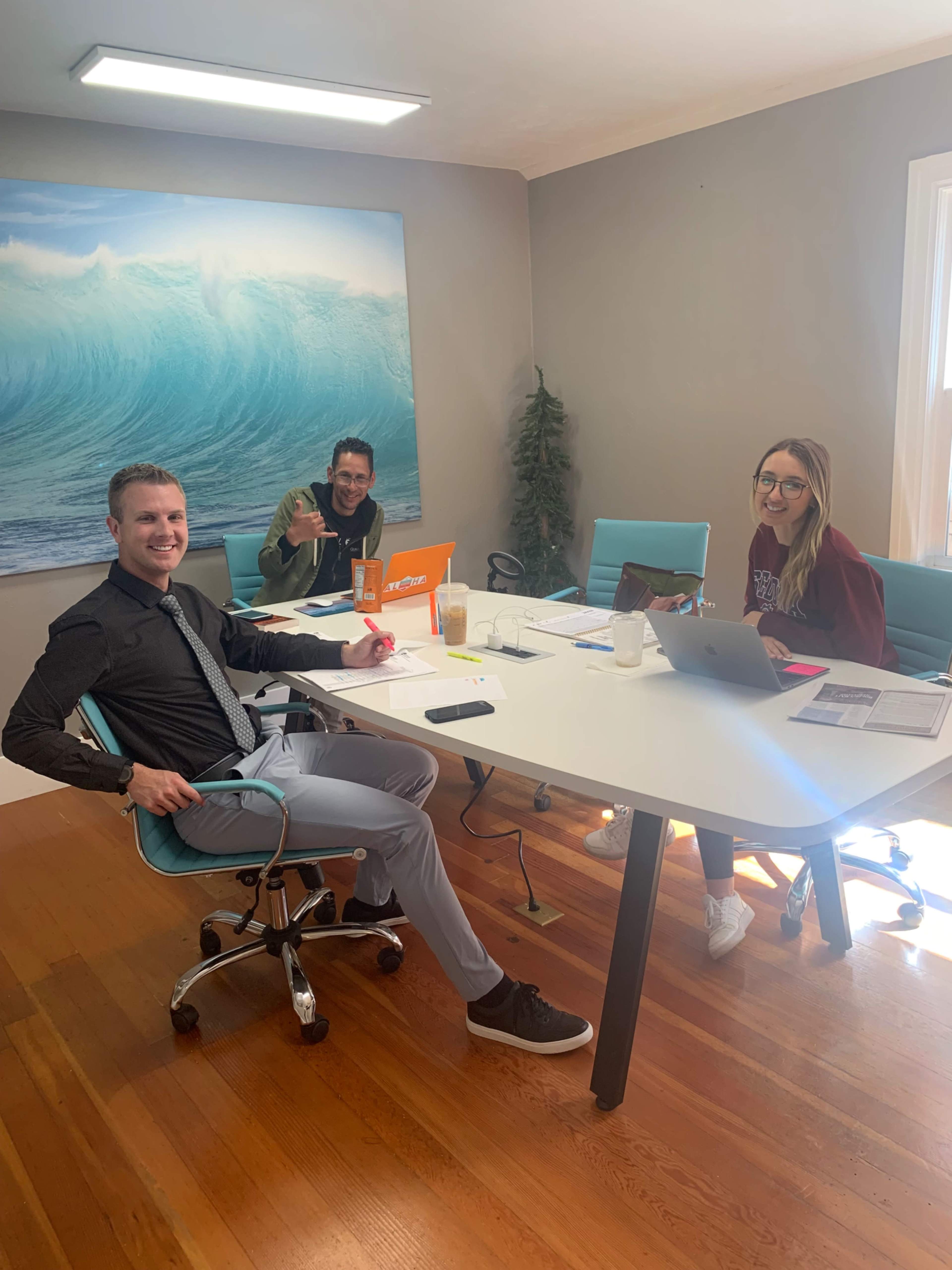 Three people are seated around a white table in a well-lit office with a large ocean wave mural on the wall.