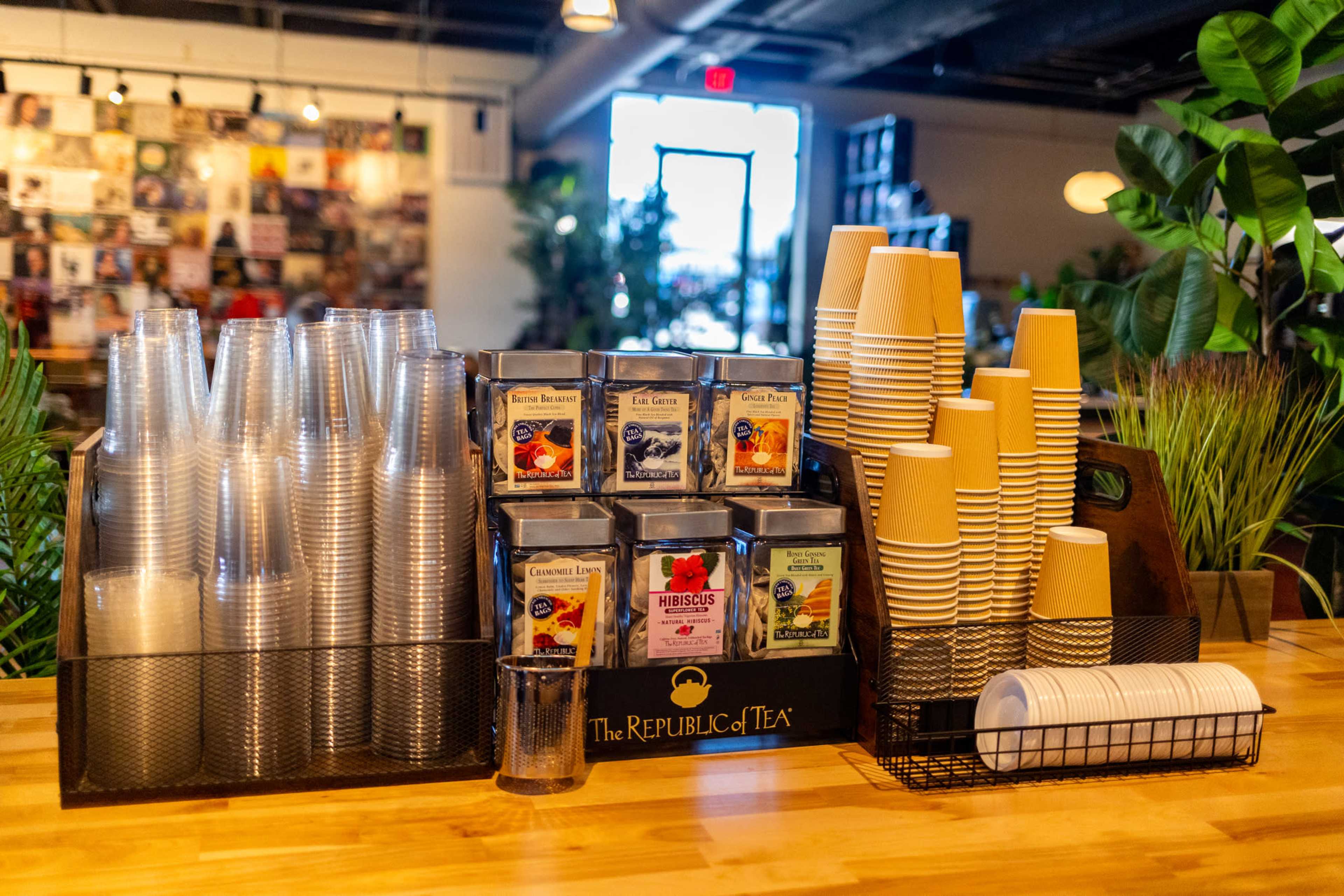 A display of tea options and disposable cups is arranged on a wooden countertop in a café.