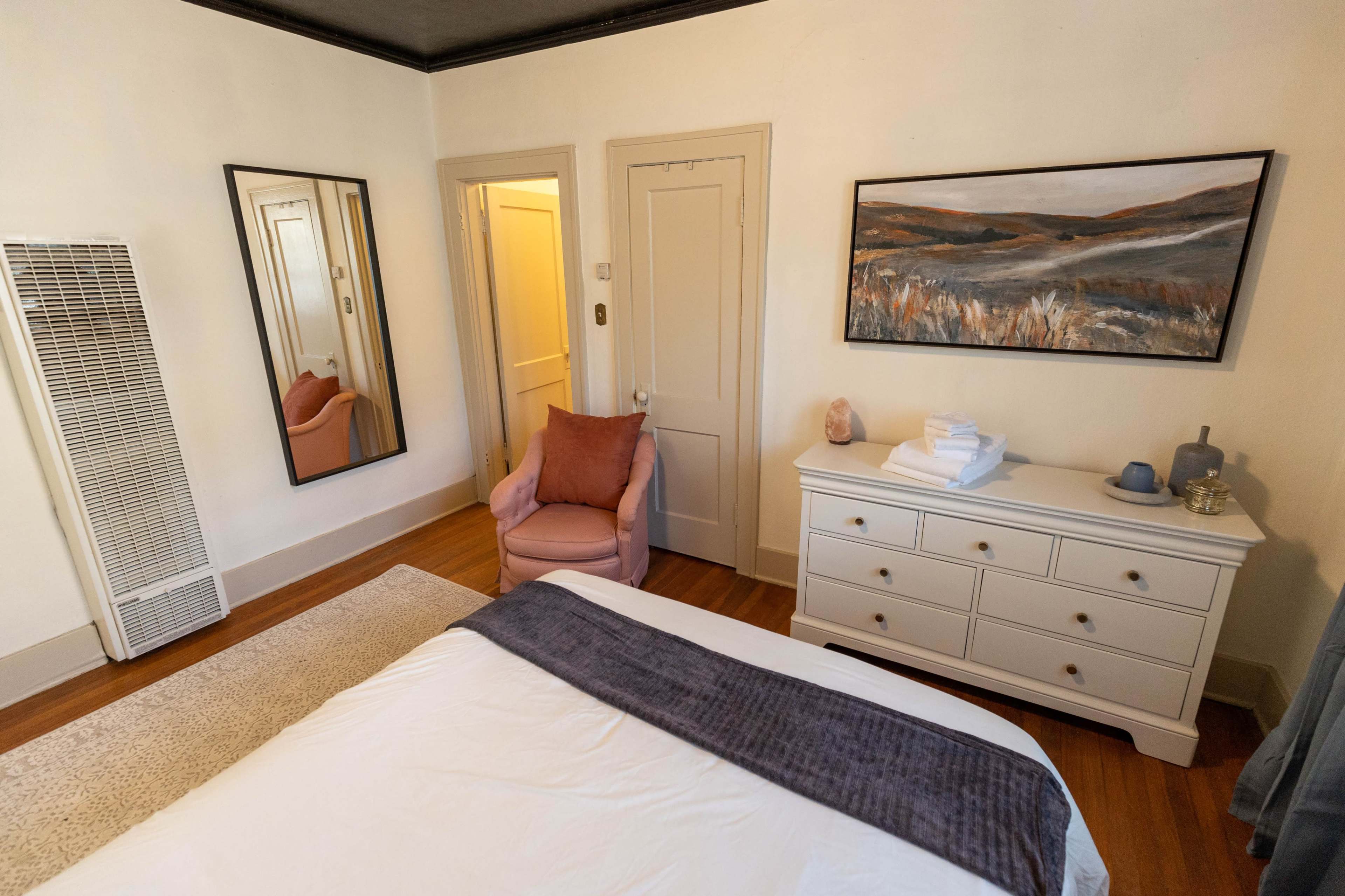 A bedroom with a bed, a dresser, a pink chair, a large mirror, and a landscape painting on the wall.