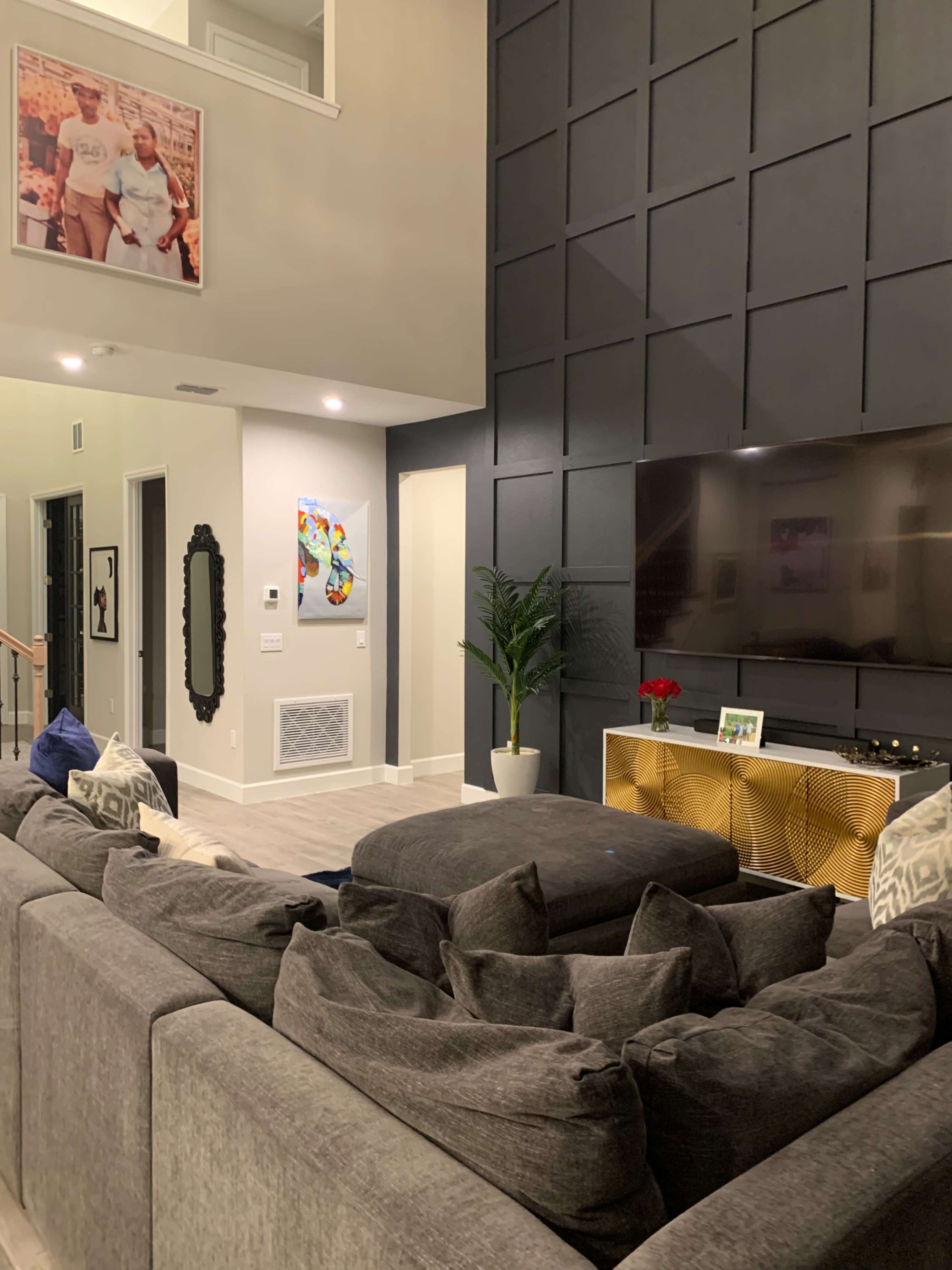 The living room features a large gray sectional sofa, a modern TV mounted on a wall with geometric paneling, and decorative elements like a plant and framed pictures.