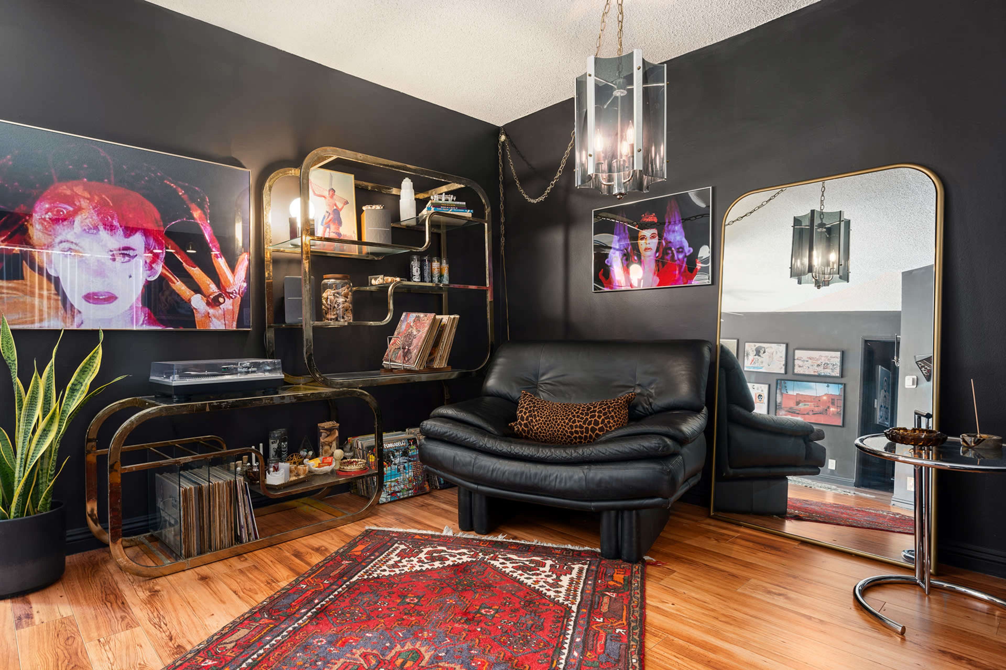 The image shows a stylish living room featuring a black leather chair, decorative shelving, a large mirror, and wall art against dark walls, with wooden flooring and a patterned rug.