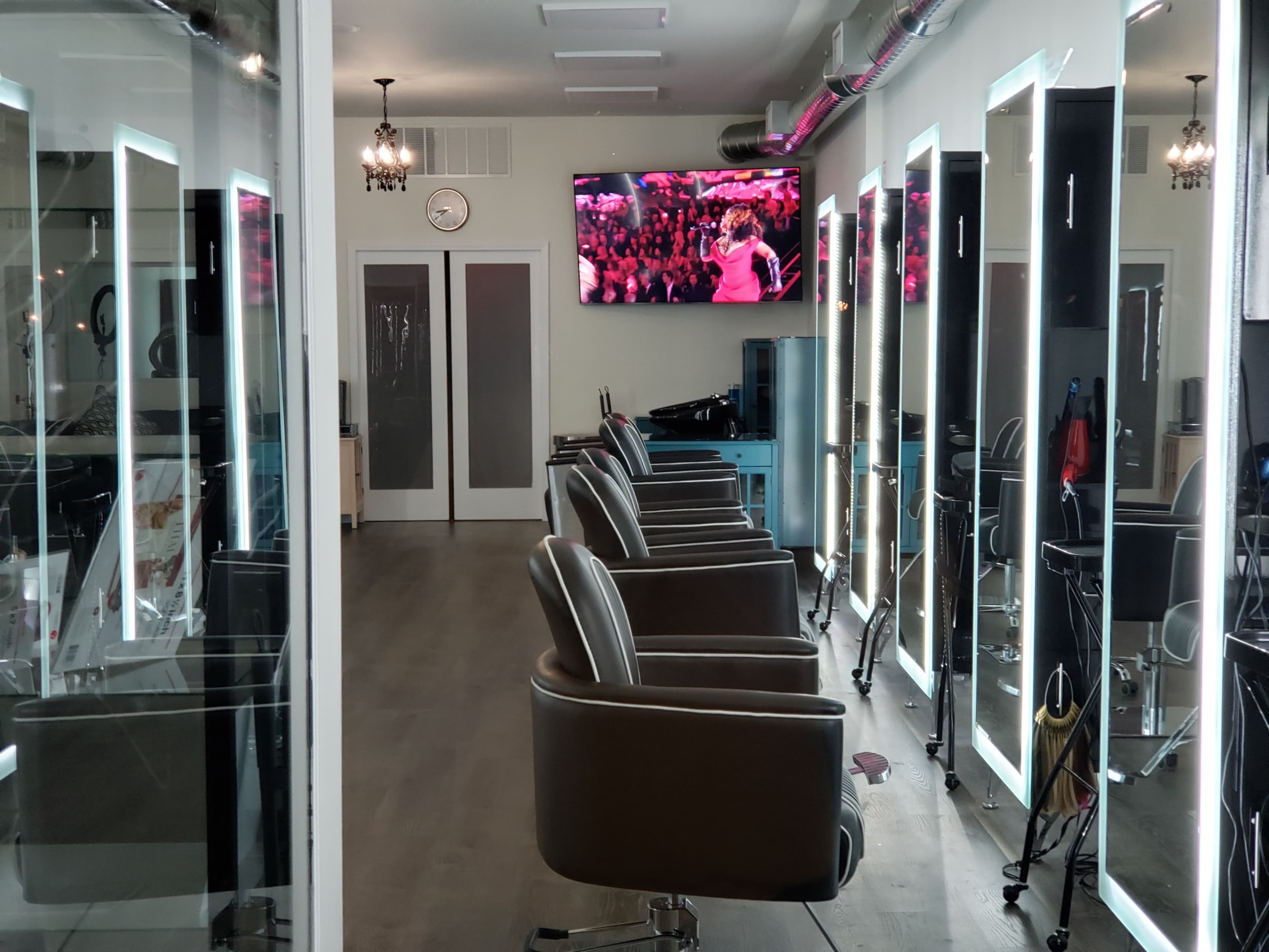 A modern hair salon with several styling chairs, illuminated mirrors, and a wall-mounted television displaying a performance.