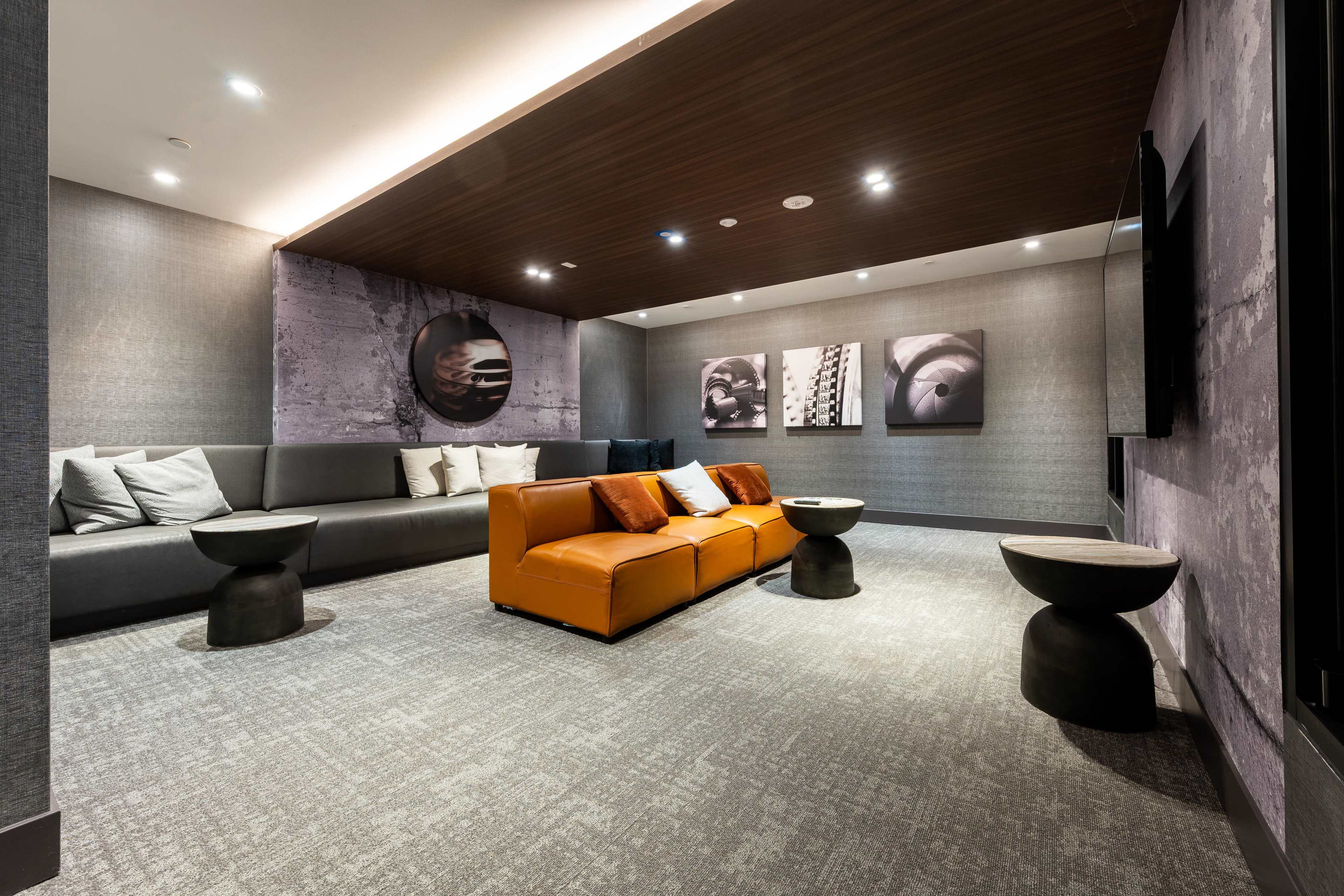 The image shows a modern lounge area featuring a gray sofa, an orange seating unit, two small black side tables, and framed artwork on the walls.