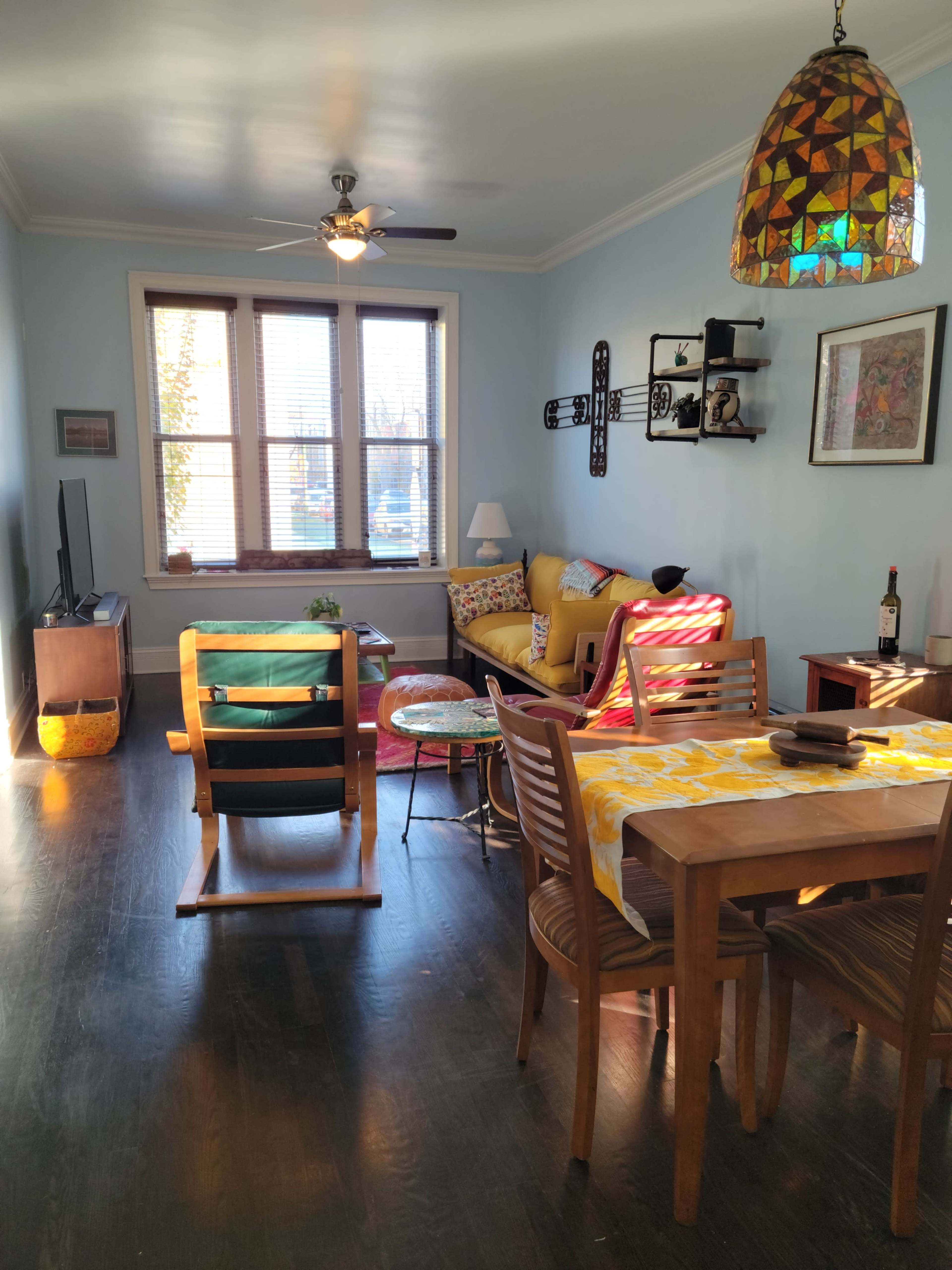 A living room with a wooden dining table, a colorful sofa, a TV, and a patterned pendant light hanging from the ceiling.