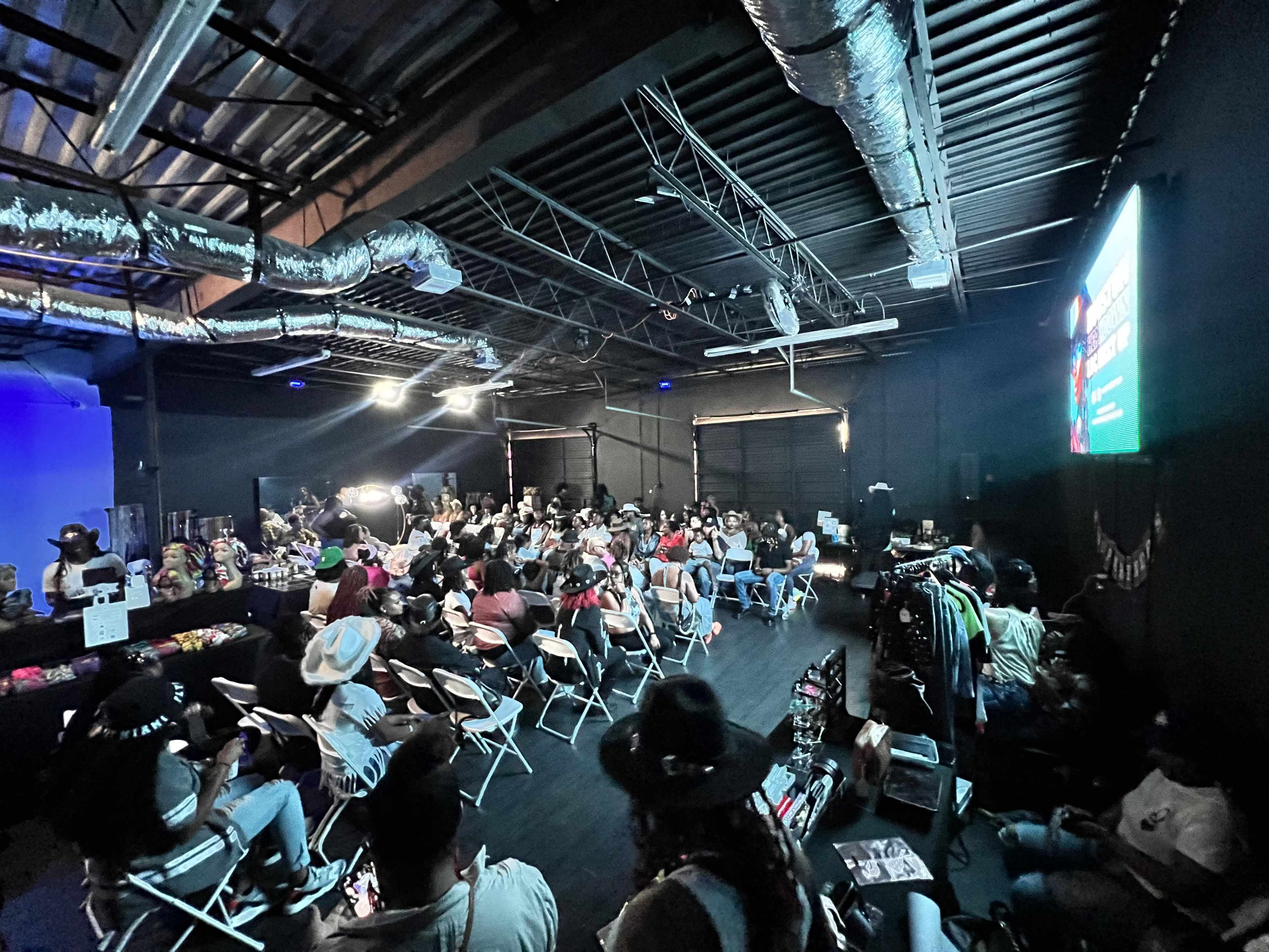 A large crowd of attendees sits in rows of folding chairs in an industrial event space, with a screen displaying graphics at the front.