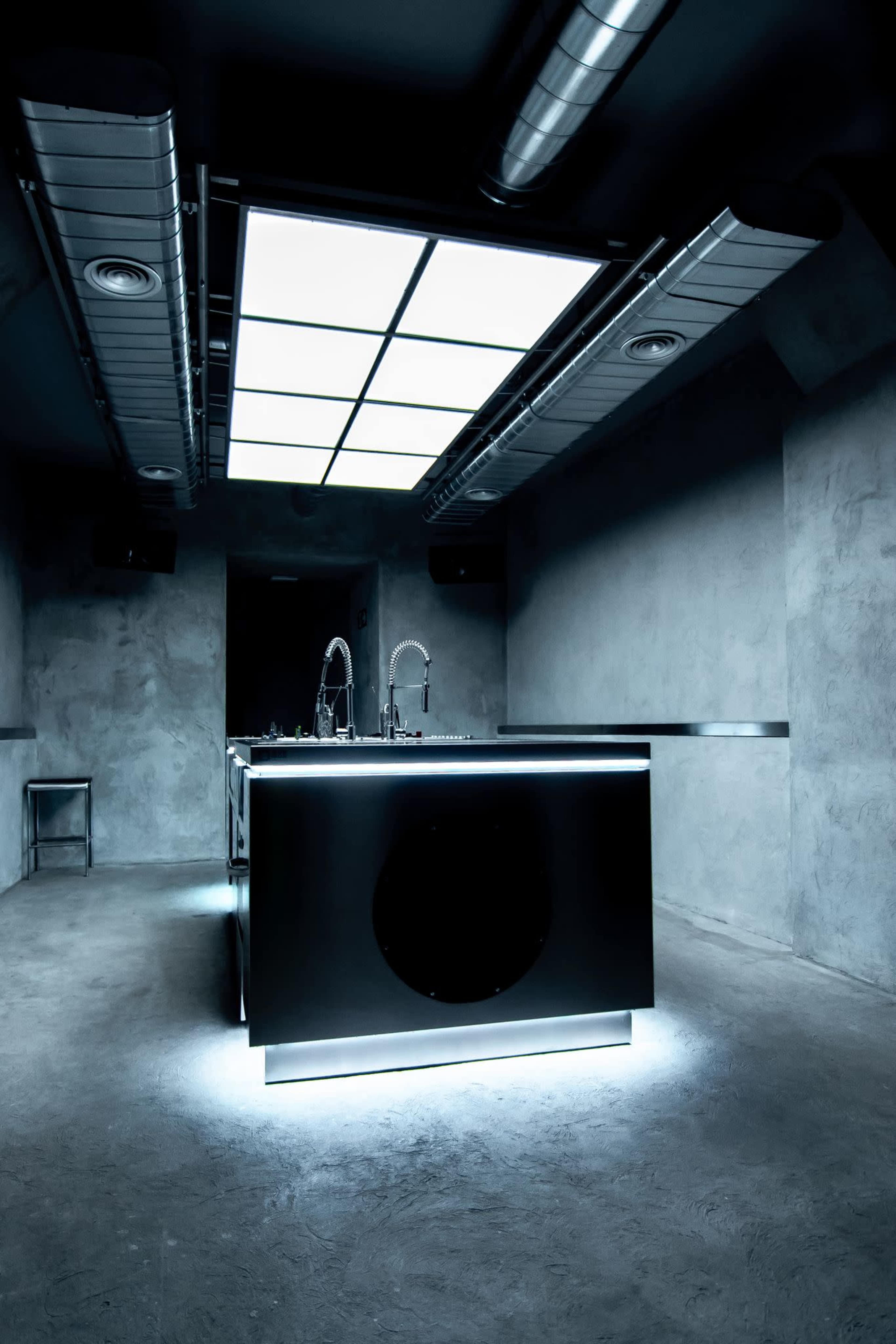 The image shows a modern kitchen with a sleek, illuminated island and industrial-style metal and concrete finishes.