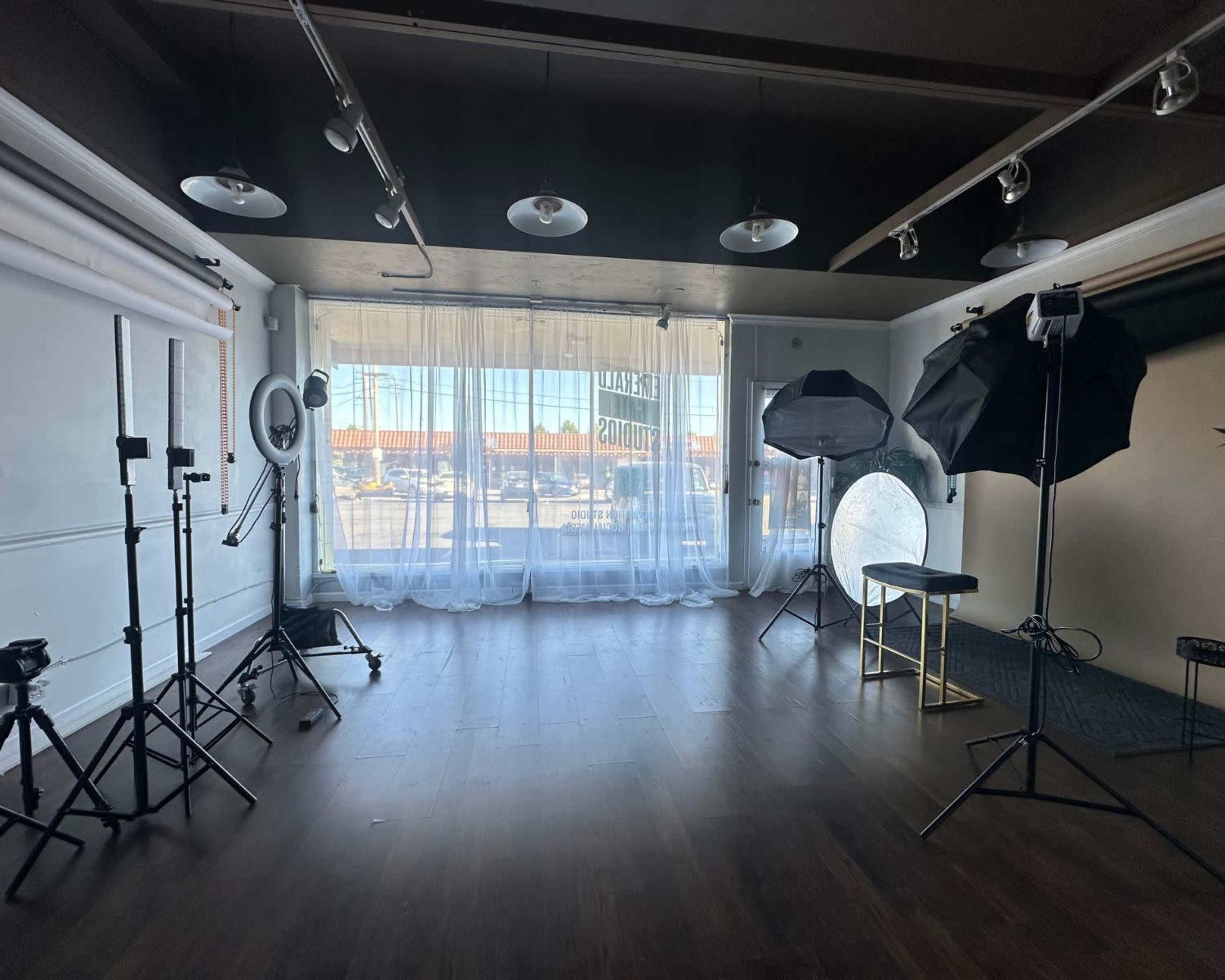 A photography studio with backdrop equipment, lighting setups, and large windows covered with sheer curtains.