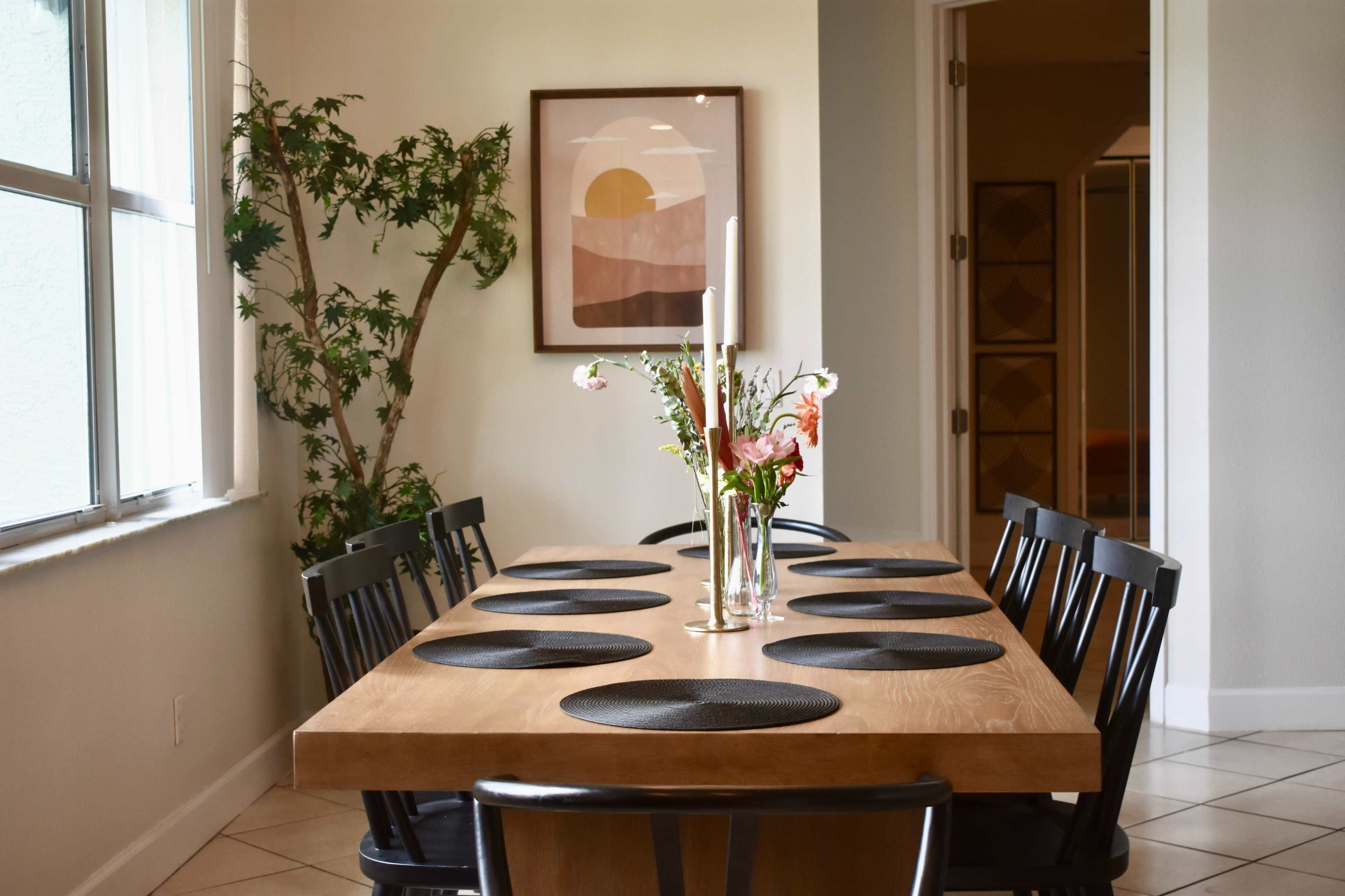 A modern dining room features a wooden table set with black placemats and a flower vase, complemented by a large artwork on the wall and a potted plant in the corner.