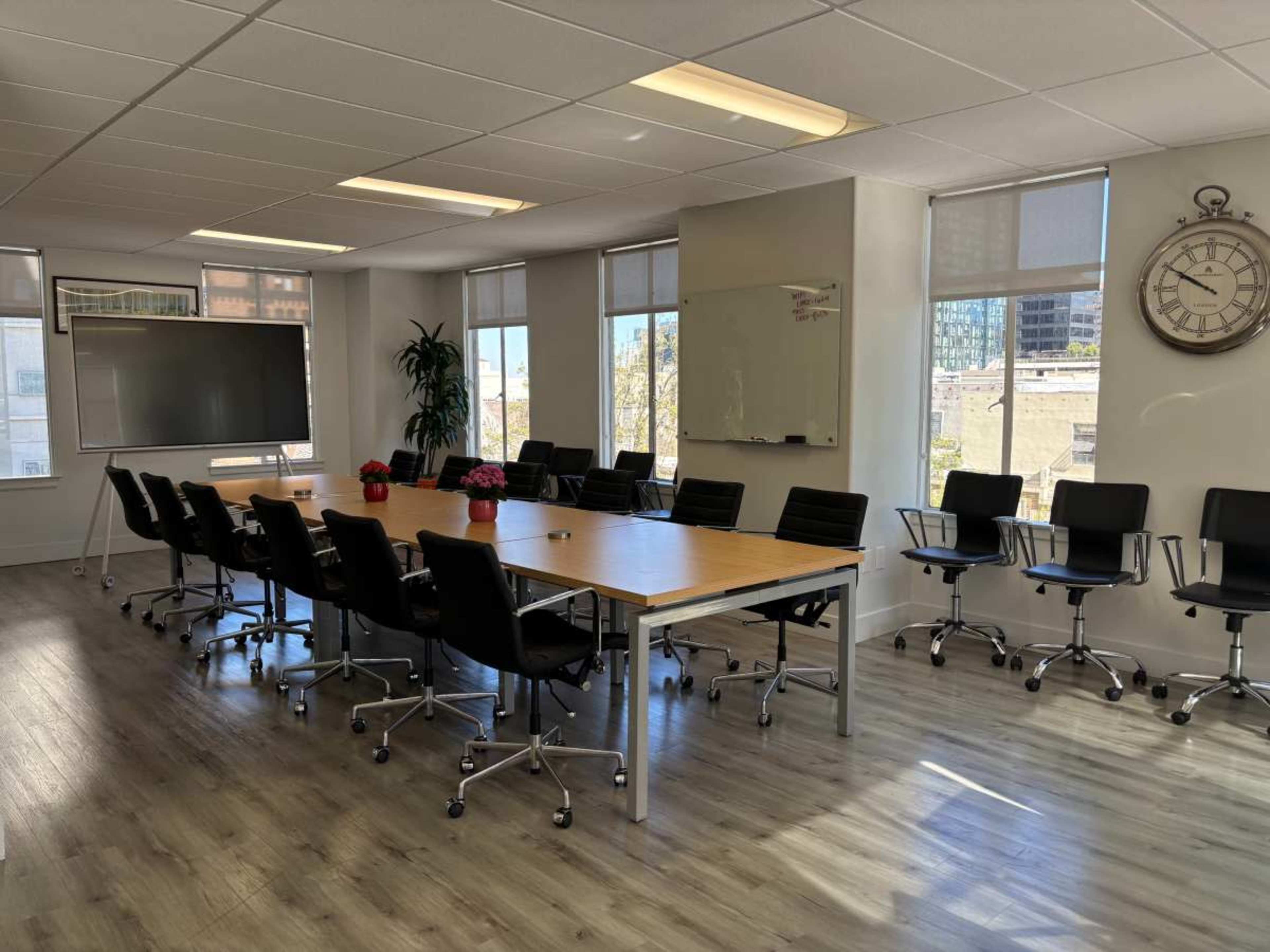 A large conference room features a long wooden table surrounded by black chairs, with large windows allowing natural light and a clock on the wall.