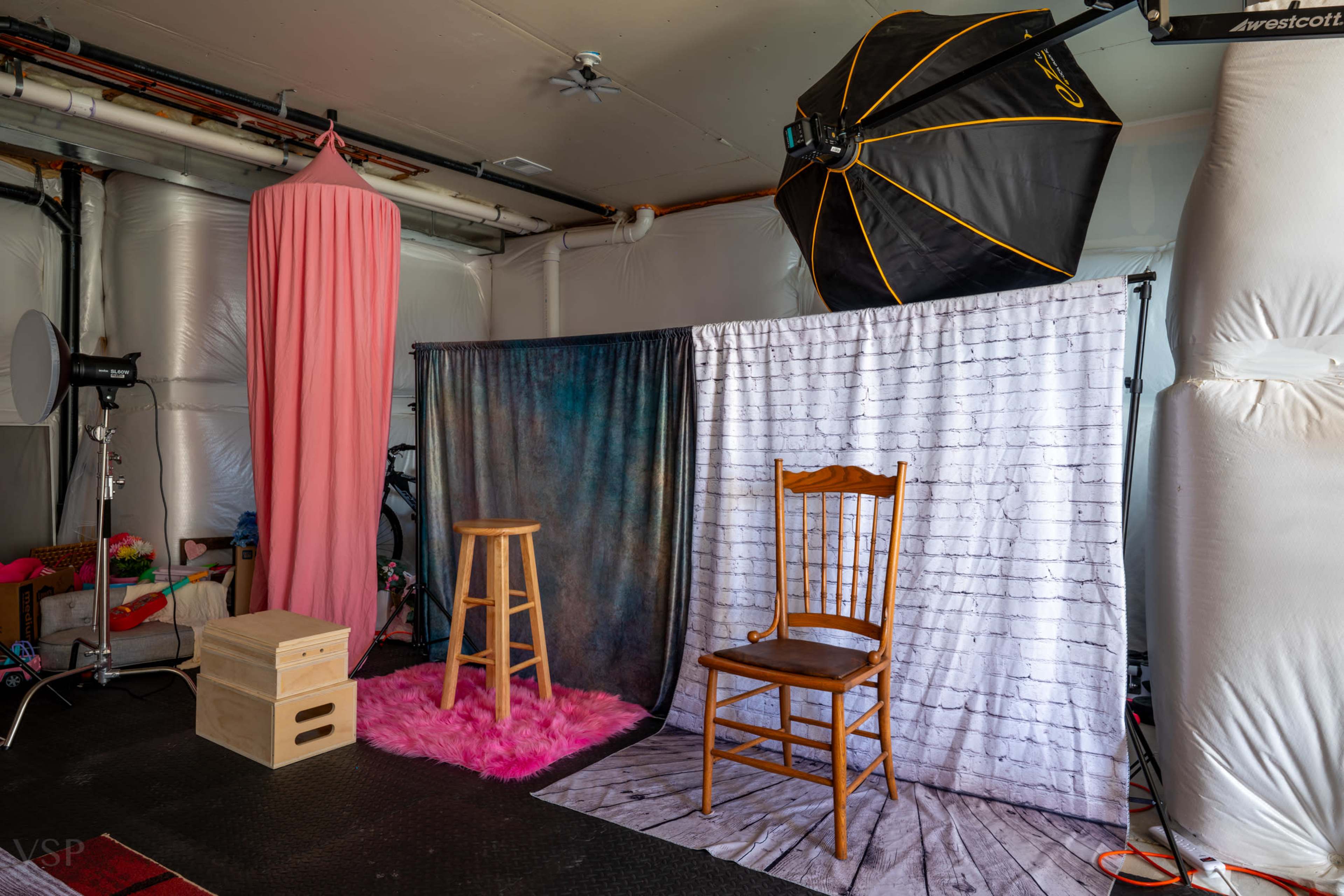 The image shows a photography studio setup with a pink draped backdrop, various colored fabric backdrops, a wooden stool, a chair, and lighting equipment.