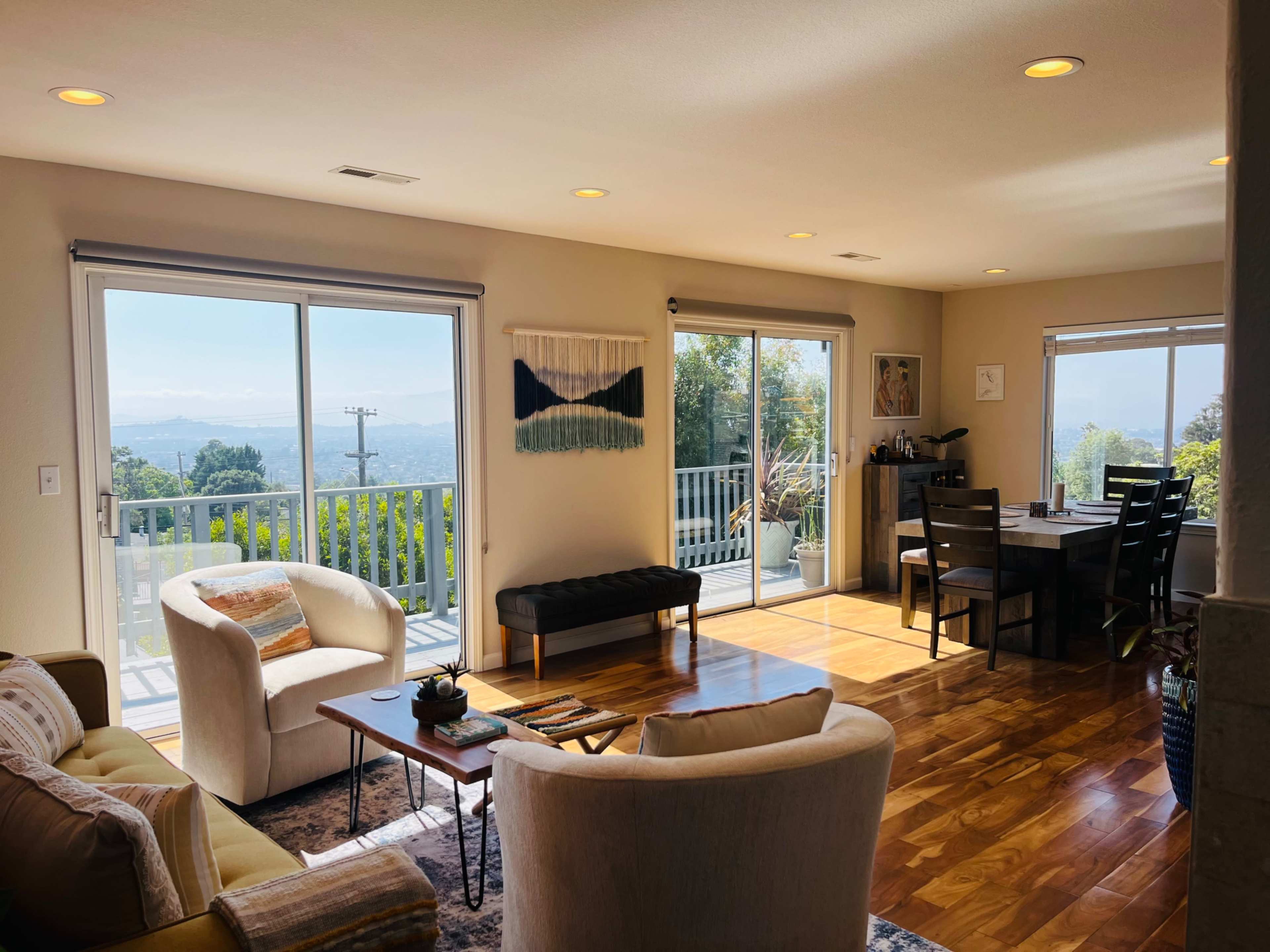 A bright living room with large windows offers a view of the outdoors, featuring contemporary furniture and a dining area nearby.