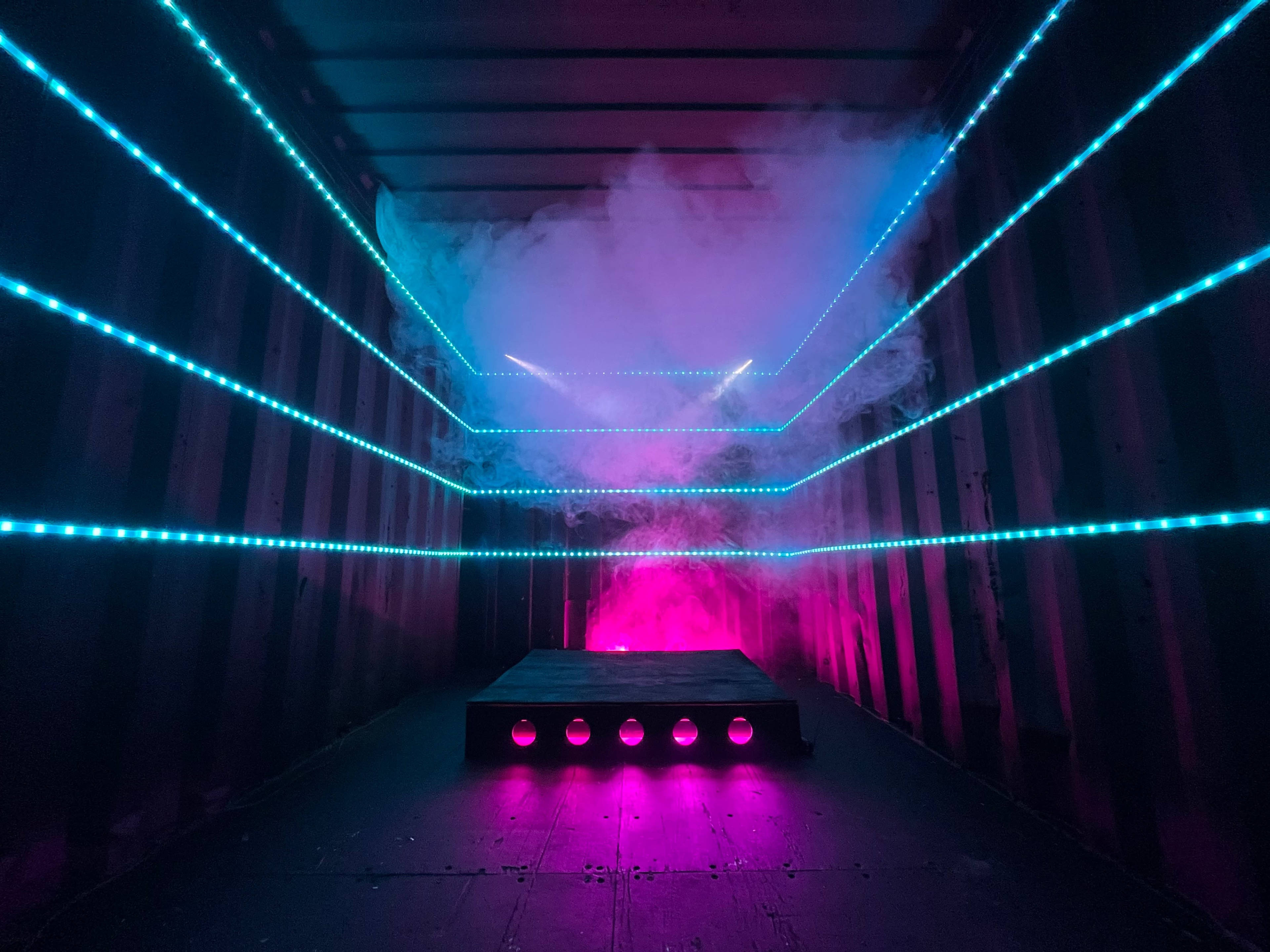 A dimly lit shipping container interior with vibrant blue and pink LED lights and fog creating a dramatic effect.