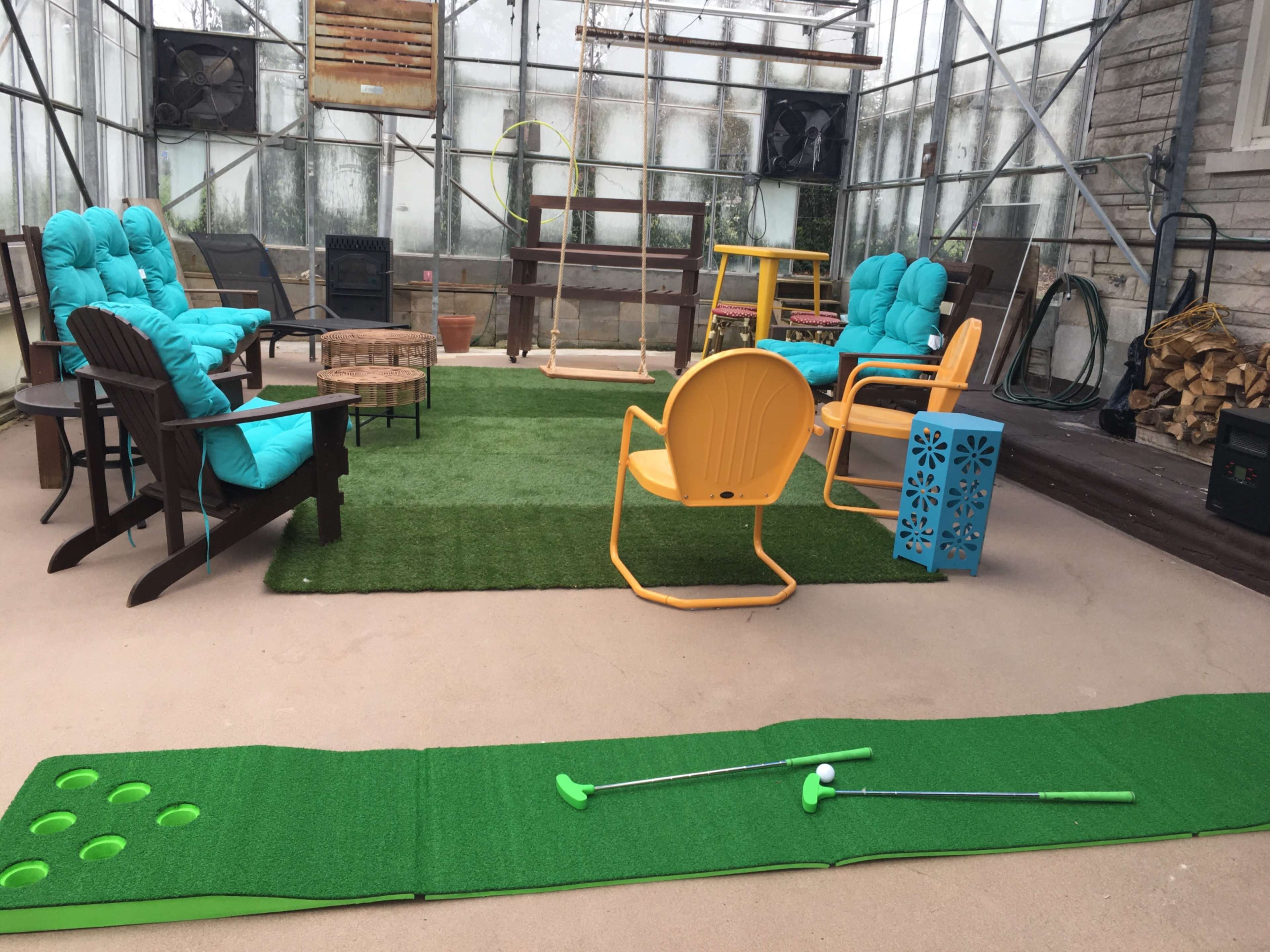 A covered outdoor area with artificial grass, various colorful chairs, and a mini golf putting green with two putters.