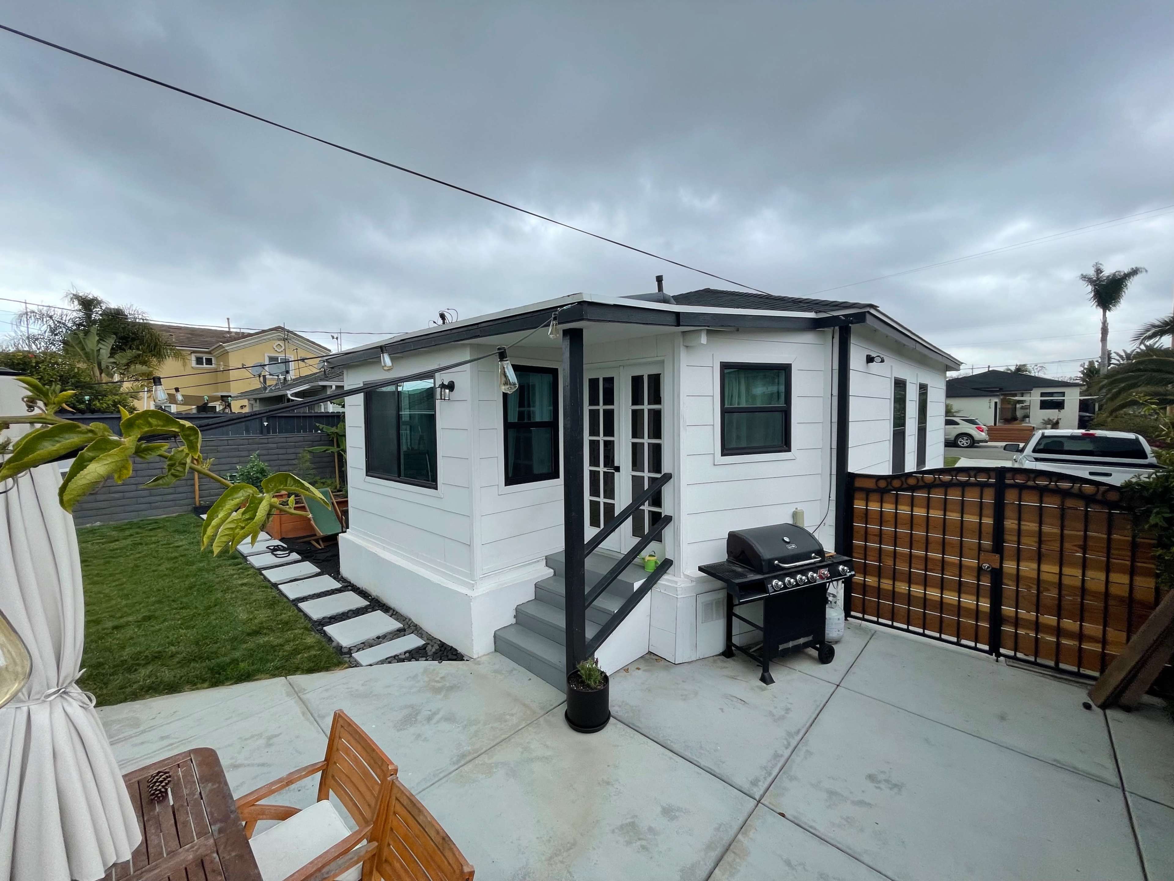 The image shows a small white house with a gray roof, a barbecue grill on a patio, and a grassy area surrounded by a wooden fence under a cloudy sky.