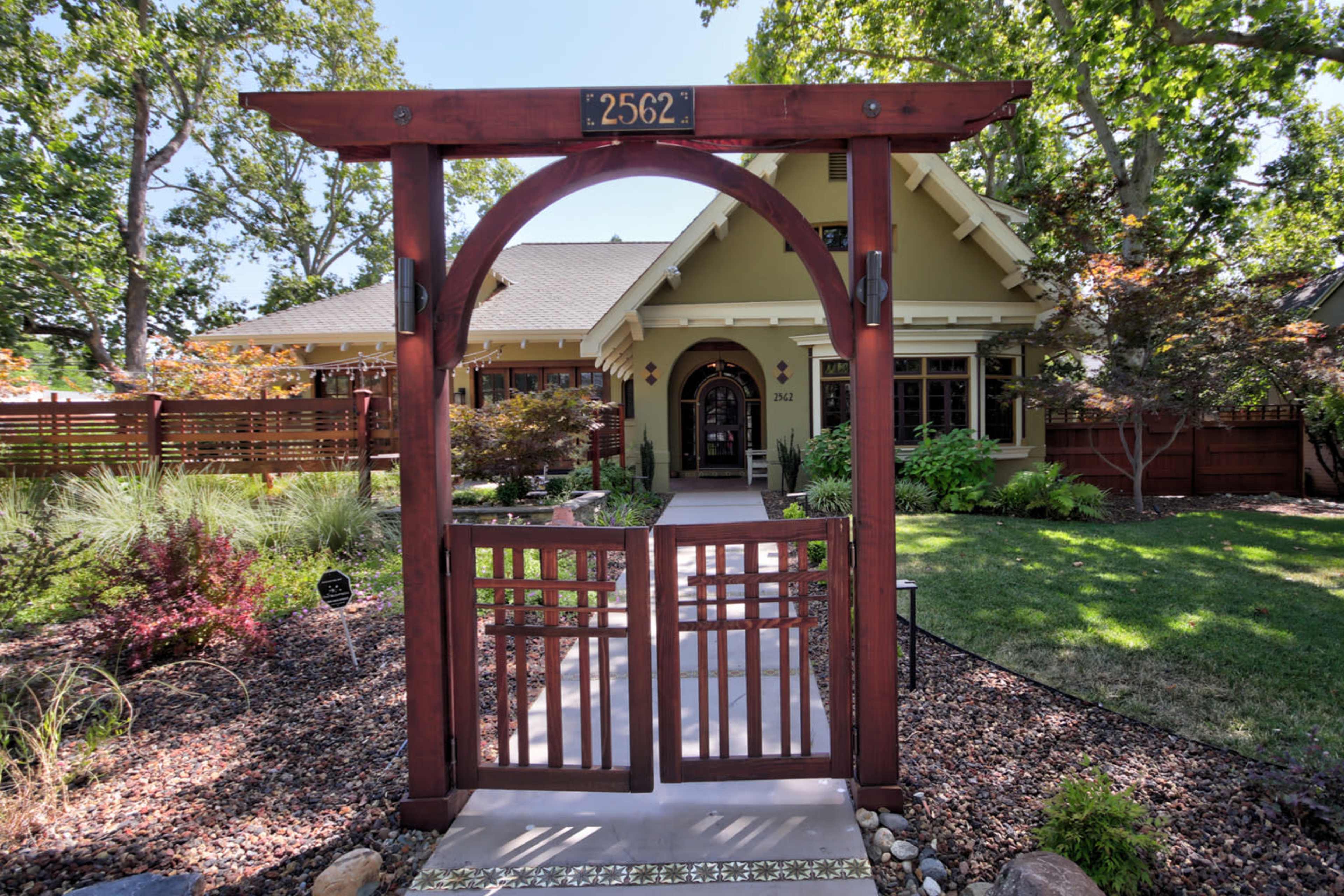 A wooden archway with a gate leads to a house surrounded by greenery and decorative landscaping.