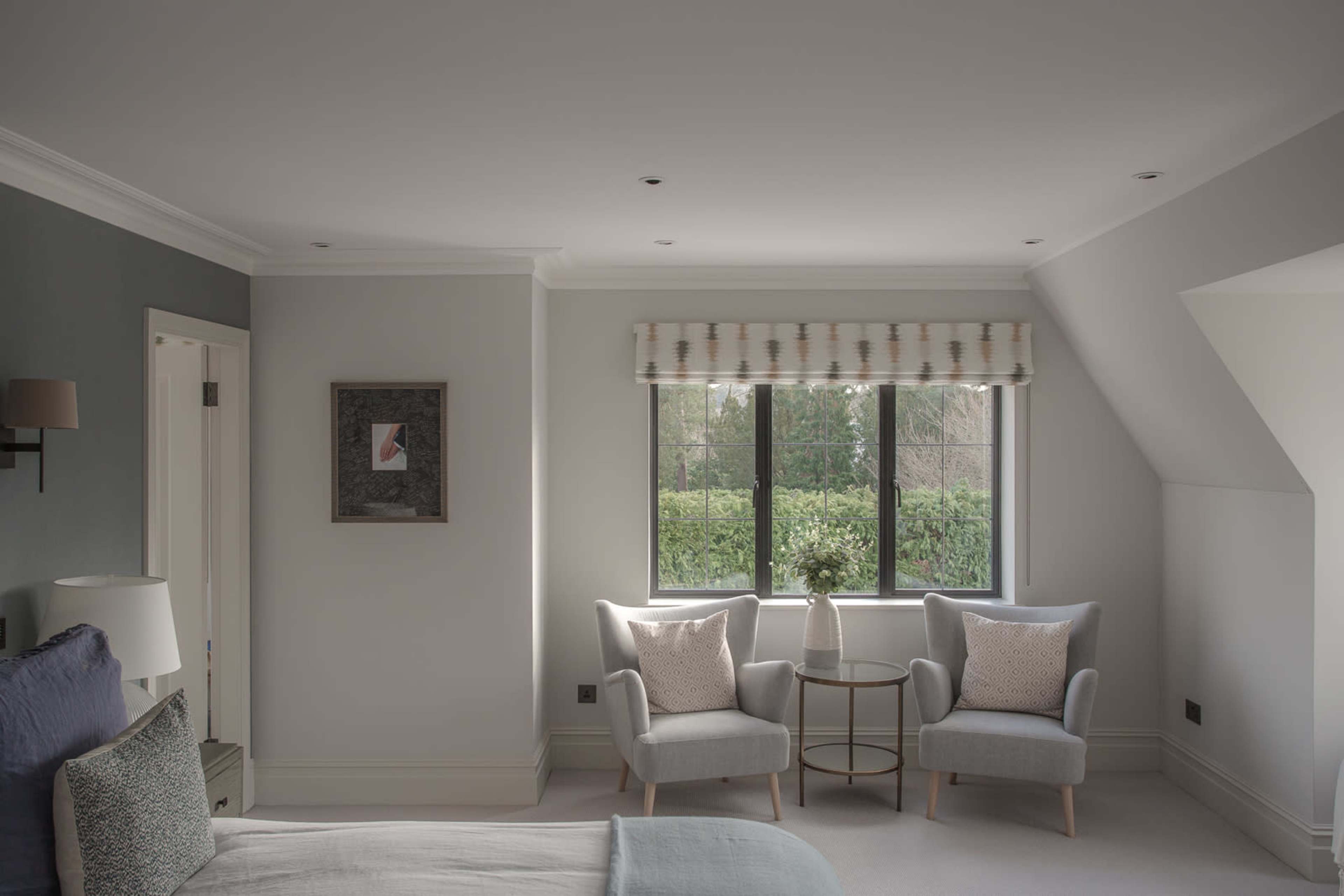 A bright, minimalist bedroom features a bed, two light gray armchairs, and a small round table near large windows overlooking greenery.