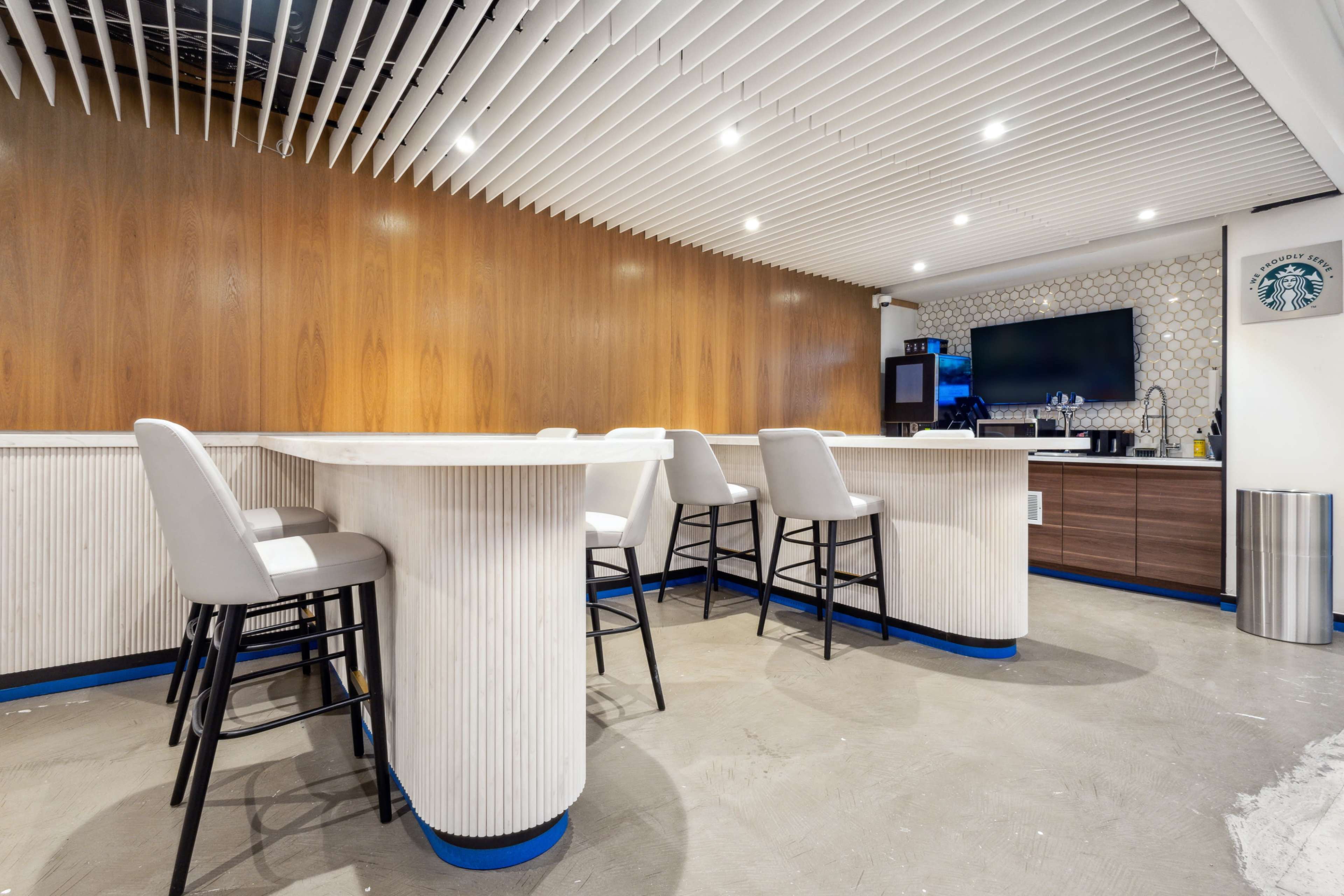 The image shows a modern bar area with four high-backed stools at a white counter, surrounded by wood paneling and illuminated by recessed lighting.