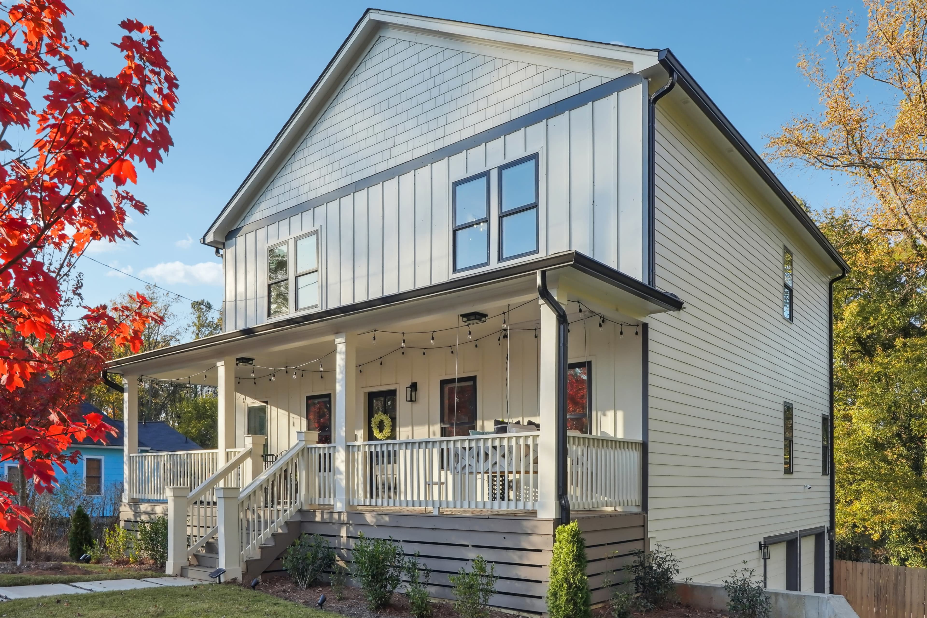 A two-story, light-colored house features a spacious front porch with string lights and landscaped greenery.