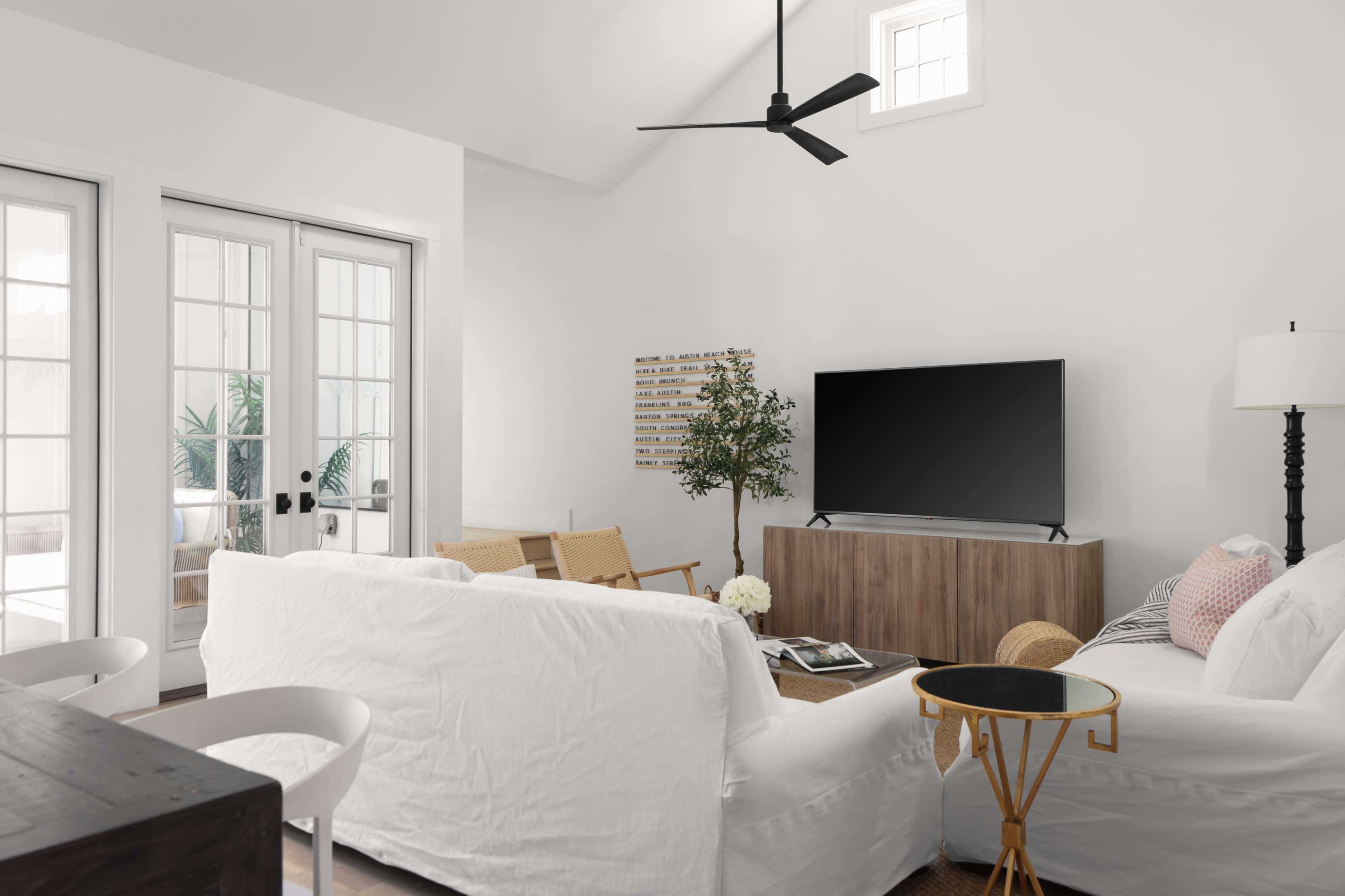 The living room features a white sectional sofa, a wooden media console with a television, and large glass doors leading to a patio with plants.
