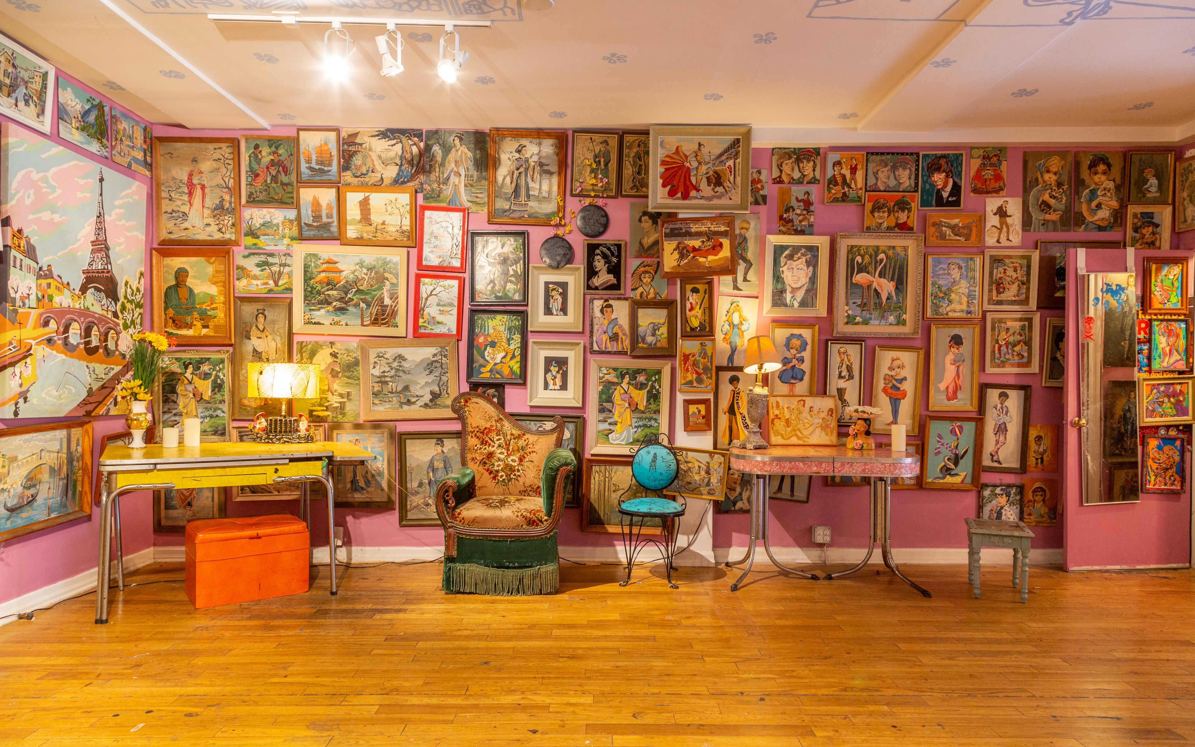 The scene features a colorful room with a wall covered in framed artwork, a vintage chair, a small table with a lamp, and a globe, all set on wooden flooring.