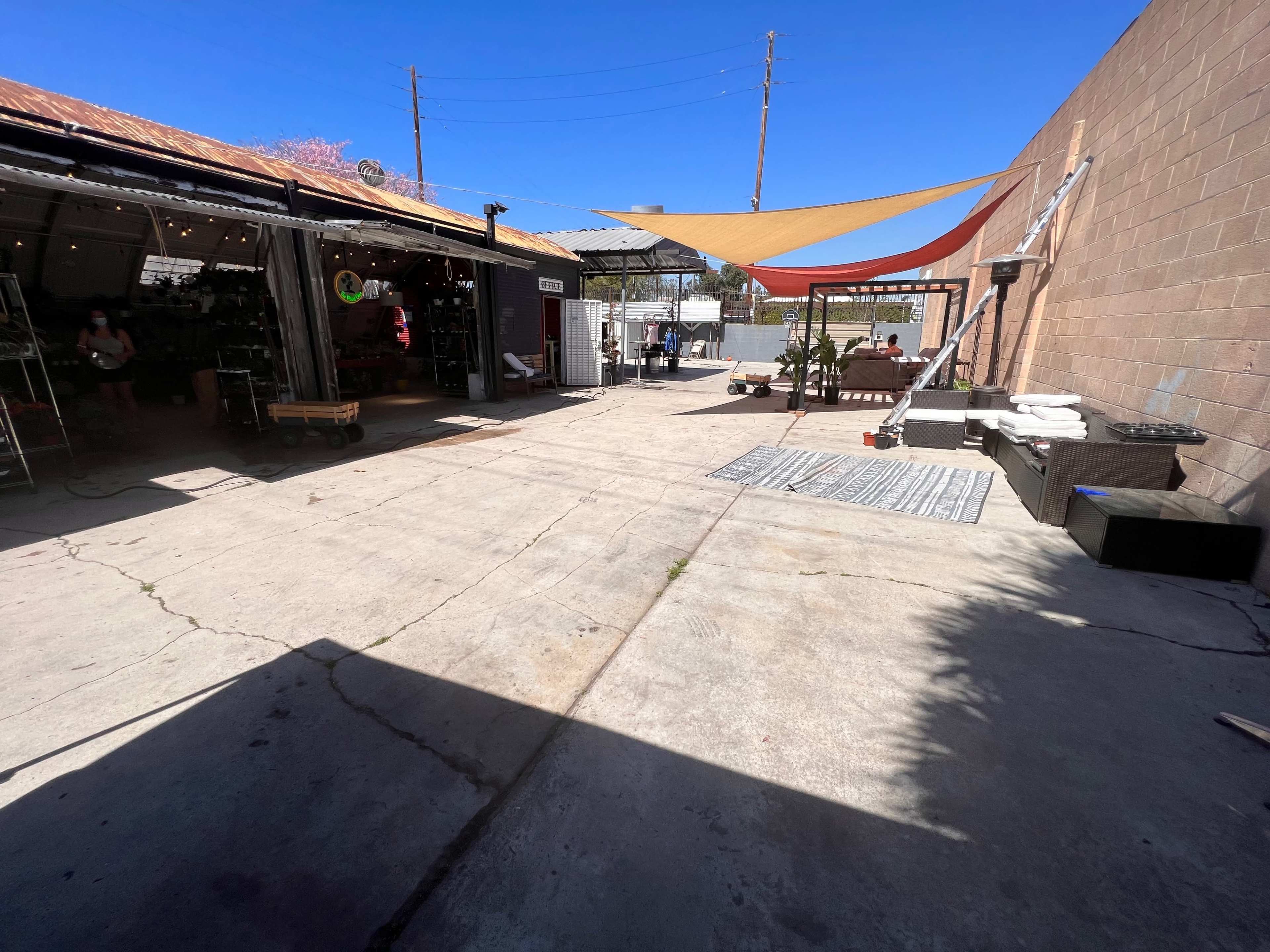 An outdoor space featuring a concrete surface, a shade structure, and various items along the walls of adjacent buildings.