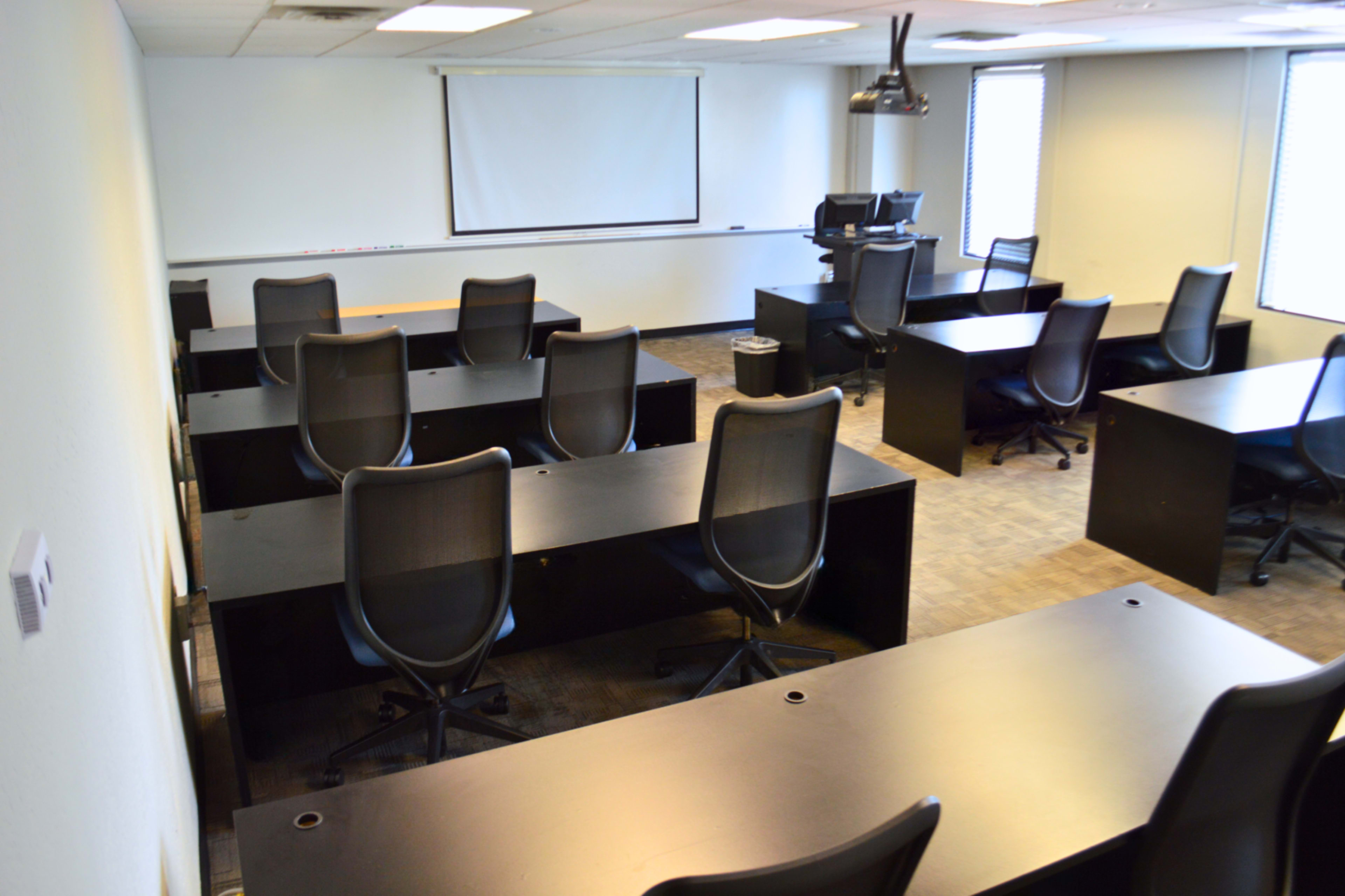 The image shows a classroom with several empty desks and chairs arranged in rows, facing a projector screen at the front.