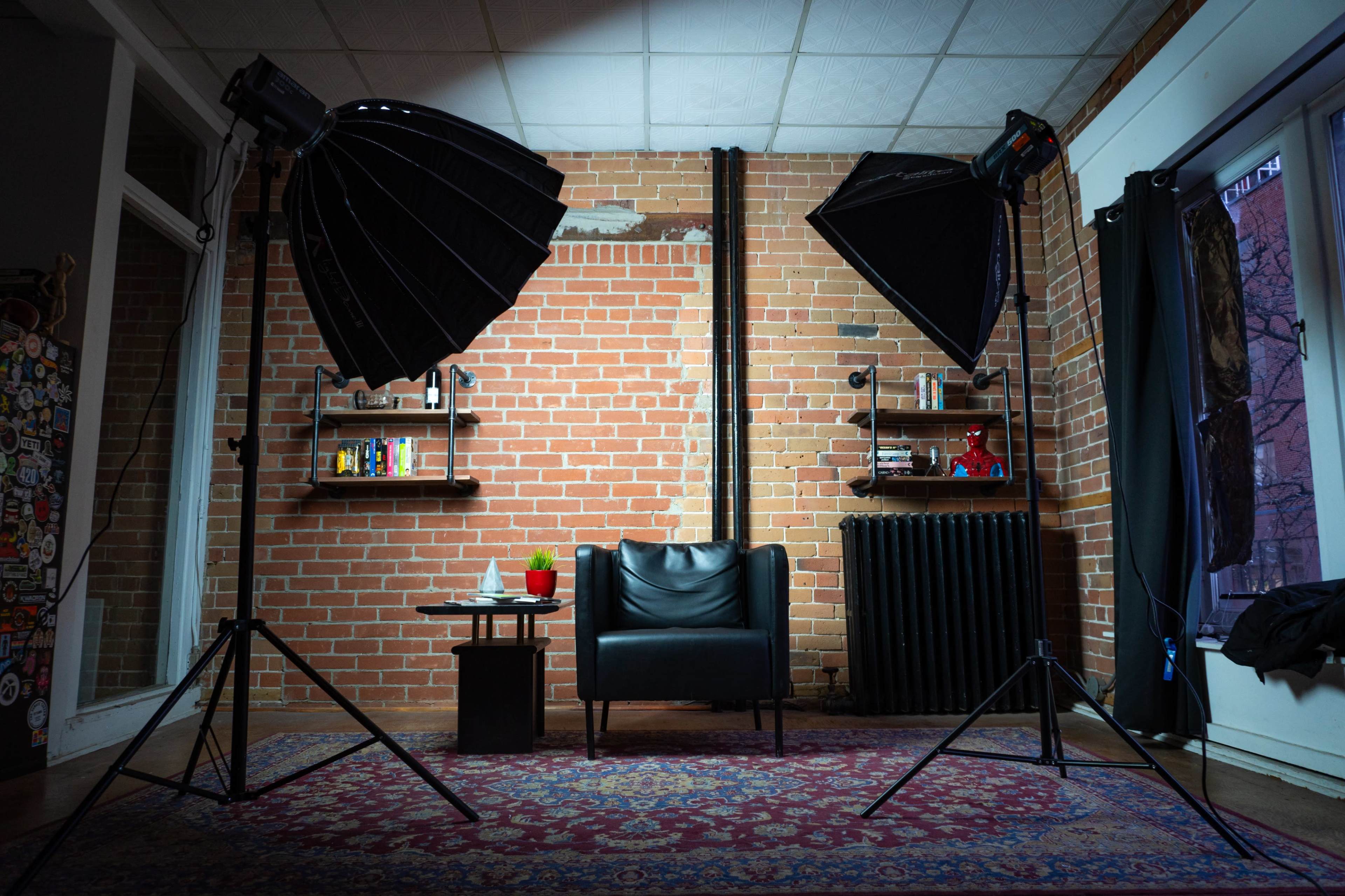 A well-lit studio setup featuring a black chair, a small table with drinks, and two softbox lights positioned on either side against a brick wall.