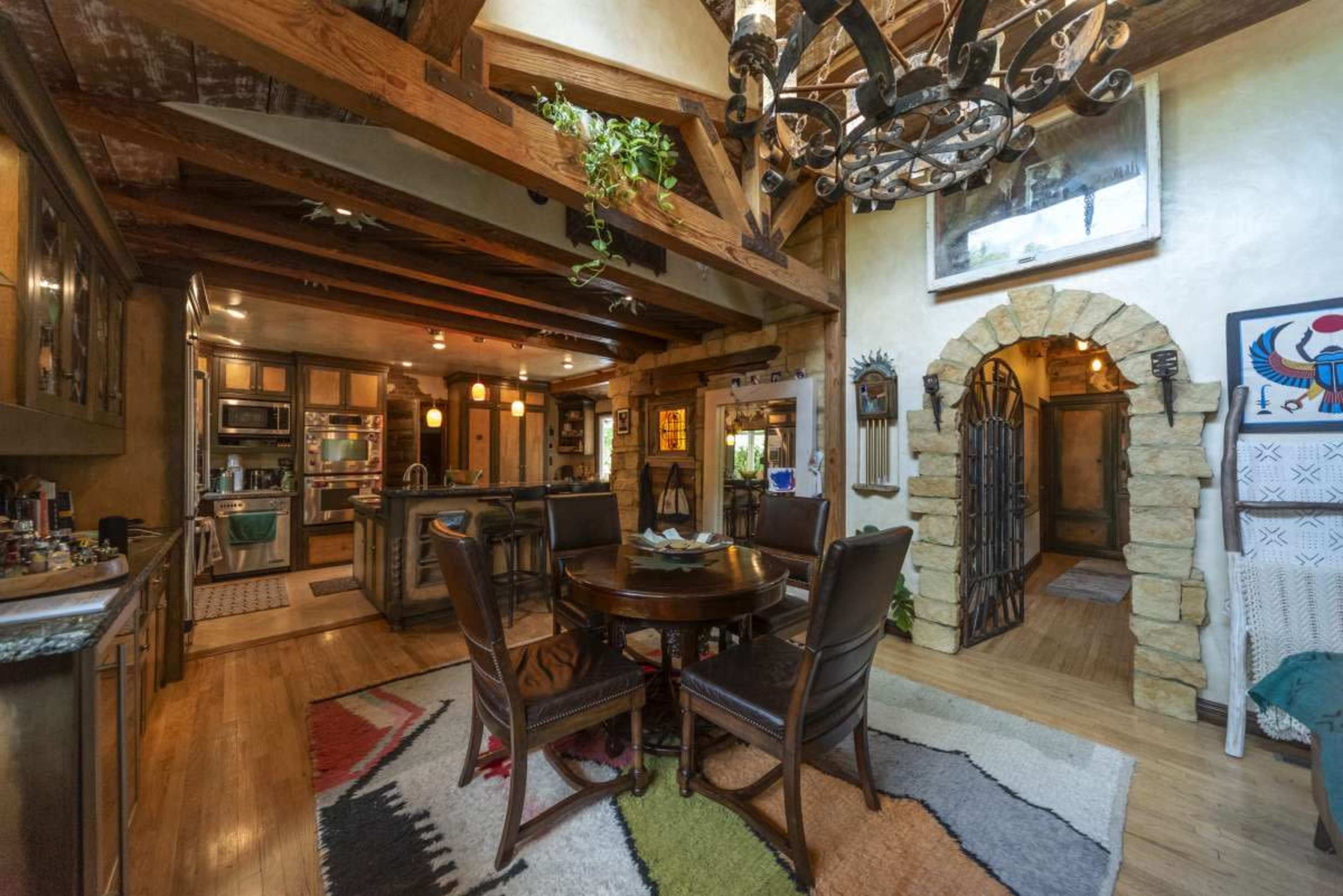 The image shows a rustic kitchen and dining area with wooden beams, a round table with four chairs, and a stone archway leading to another room.