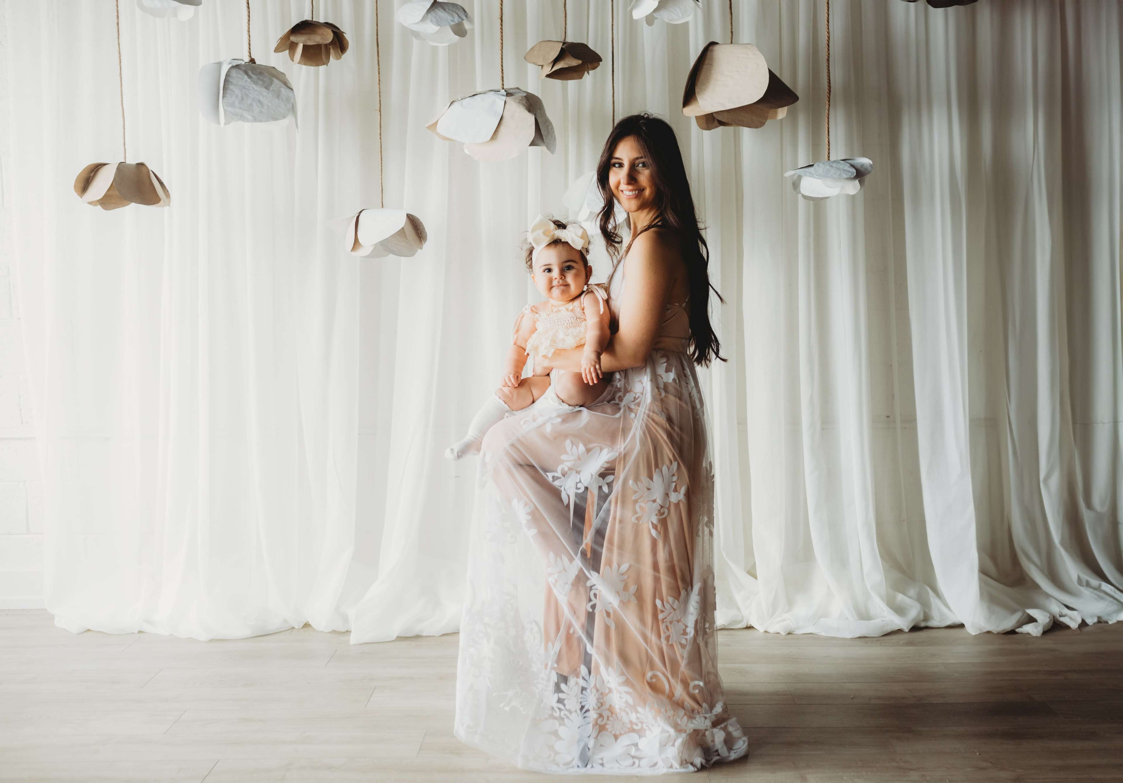 A woman holds a young girl as they stand against a backdrop of hanging paper clouds and draped fabric.