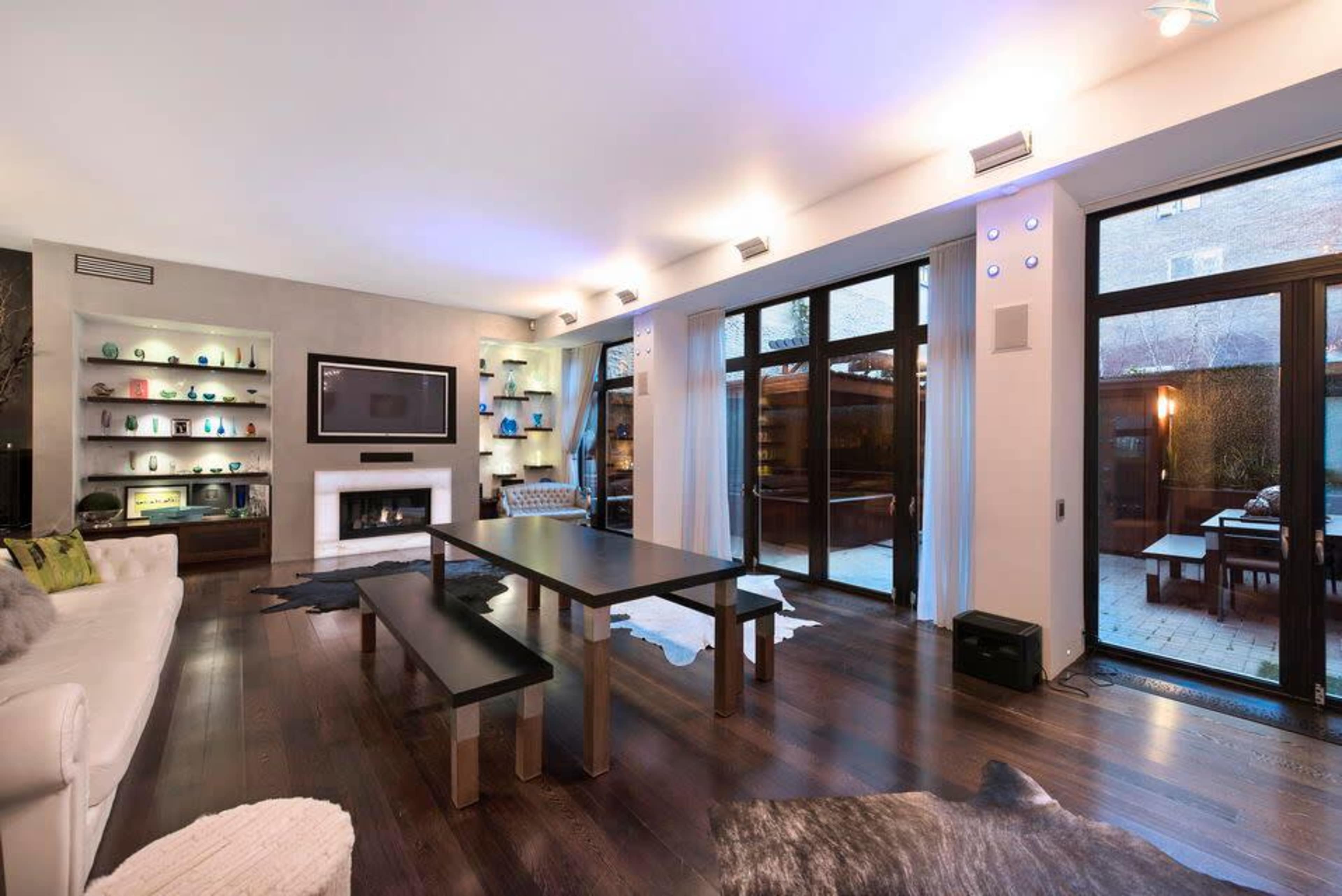 A modern living room with large windows, a flat-screen TV mounted on the wall, and a wooden table set on a dark hardwood floor.