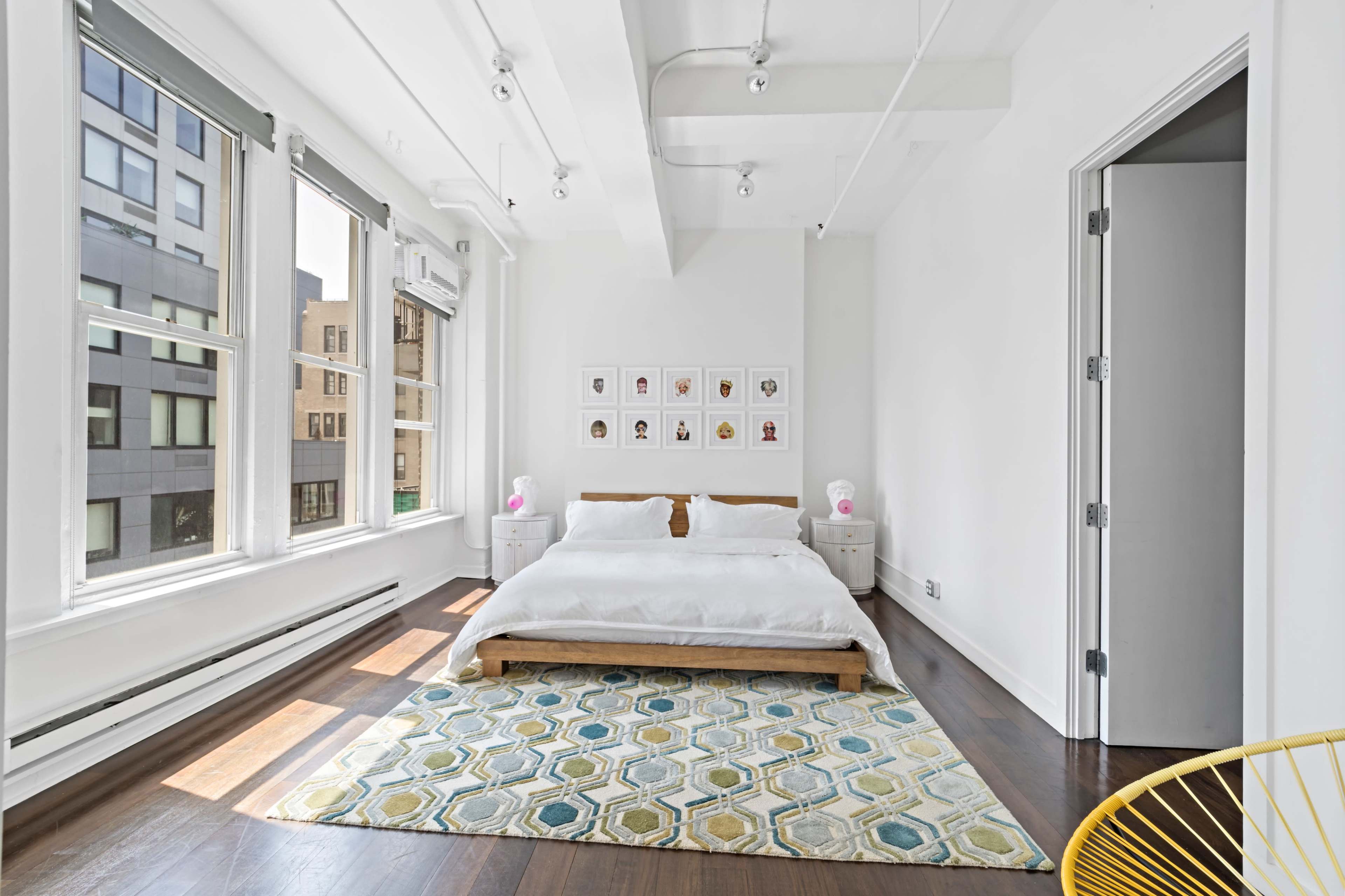 A bright bedroom features a large bed with white bedding, a patterned rug, and windows that provide natural light.