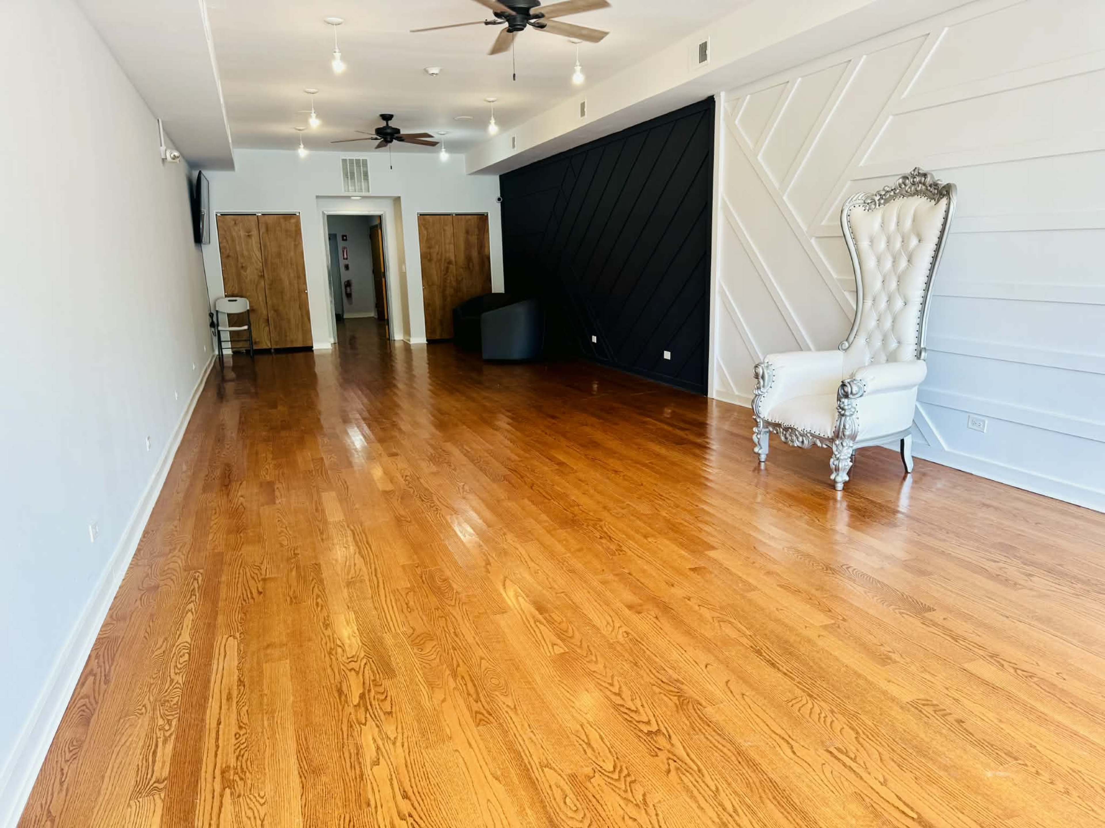The image shows a spacious room with polished wooden floors, a white wall with a patterned design, a black accent wall, and a large ornate white chair in one corner.