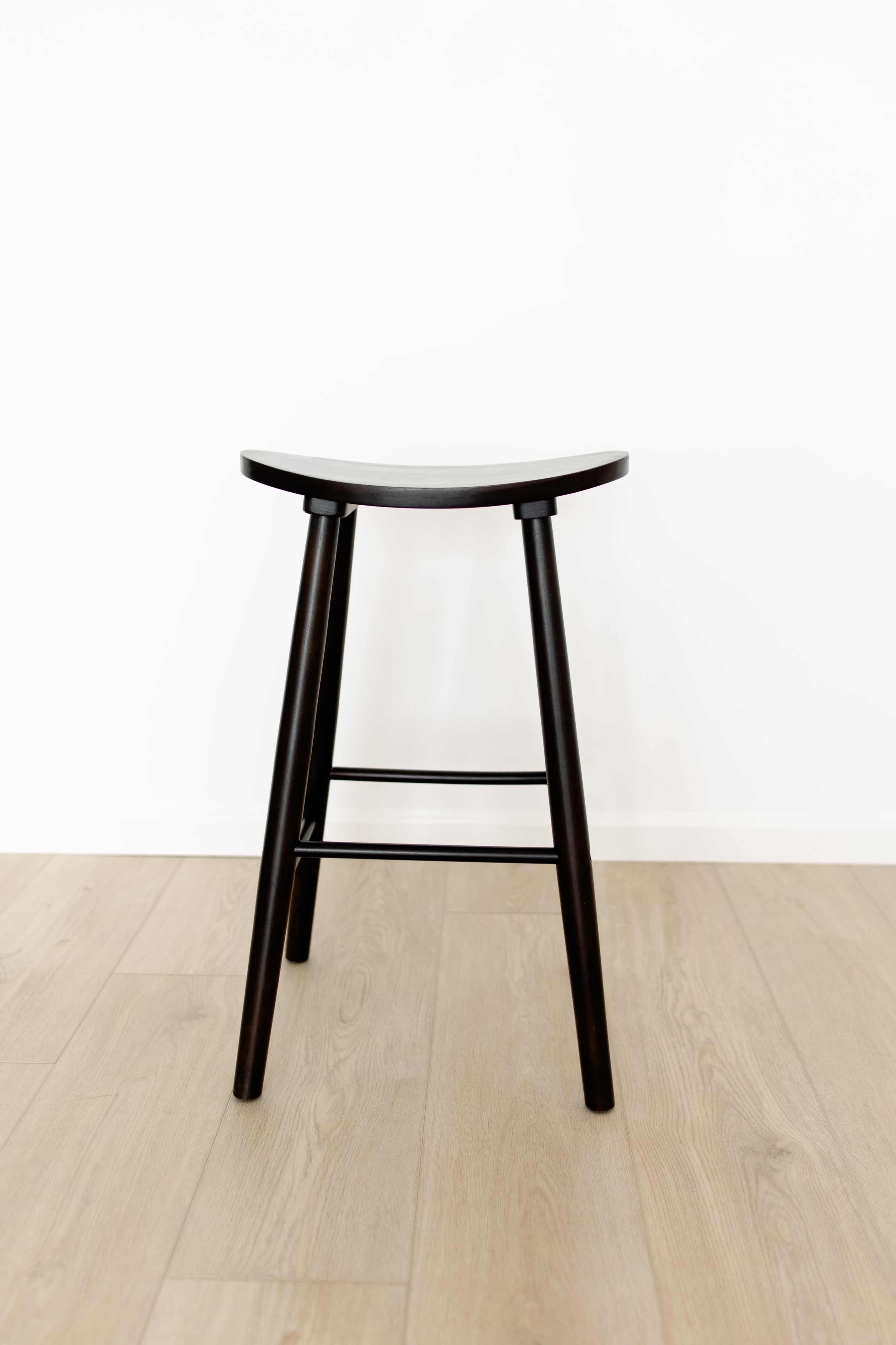 A black wooden stool with a curved seat and four legs sits on a light wood floor against a white wall.
