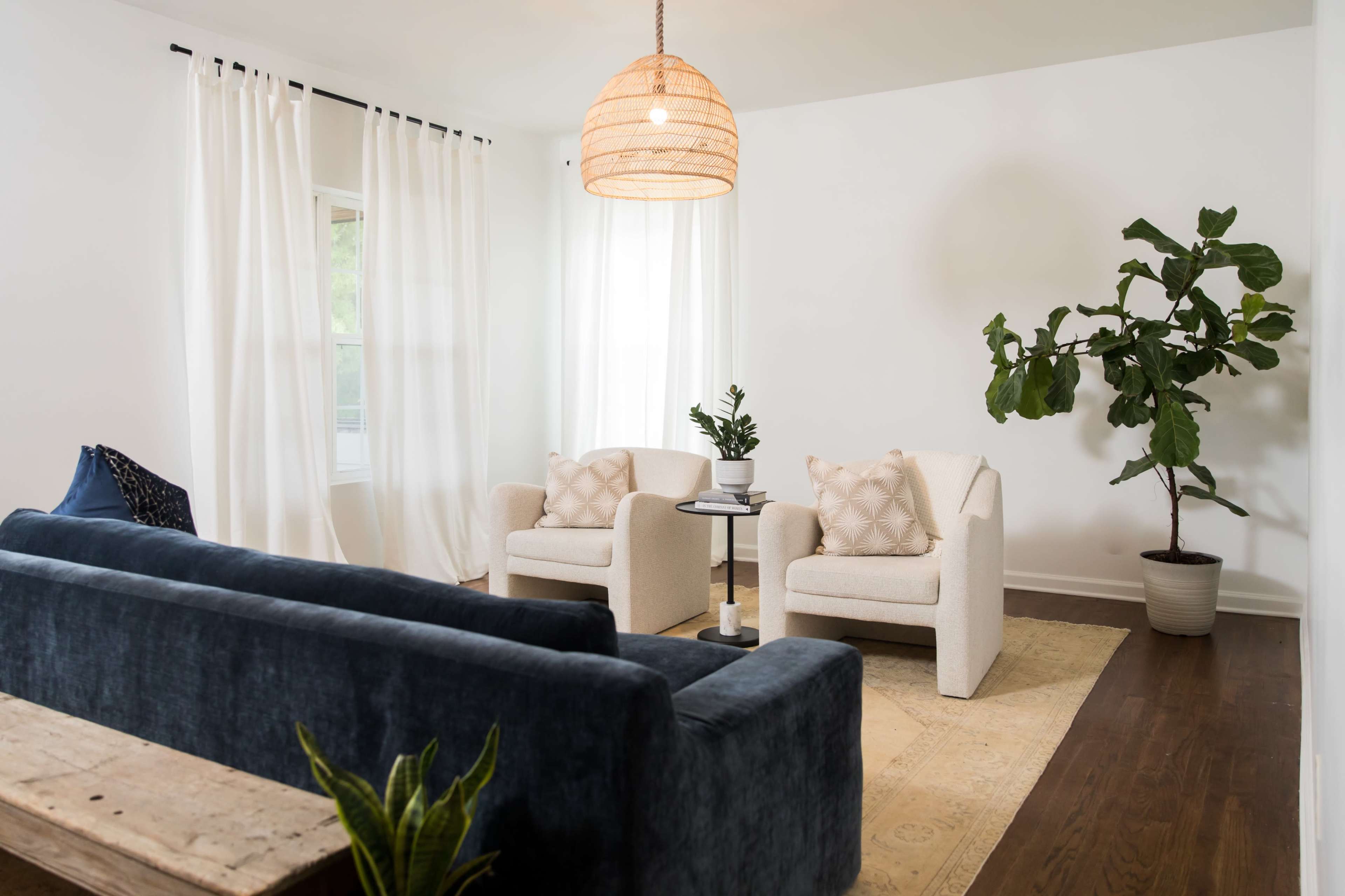 A modern living room featuring a dark blue sofa, two light-colored armchairs, a coffee table, a potted plant, and large windows with white curtains.