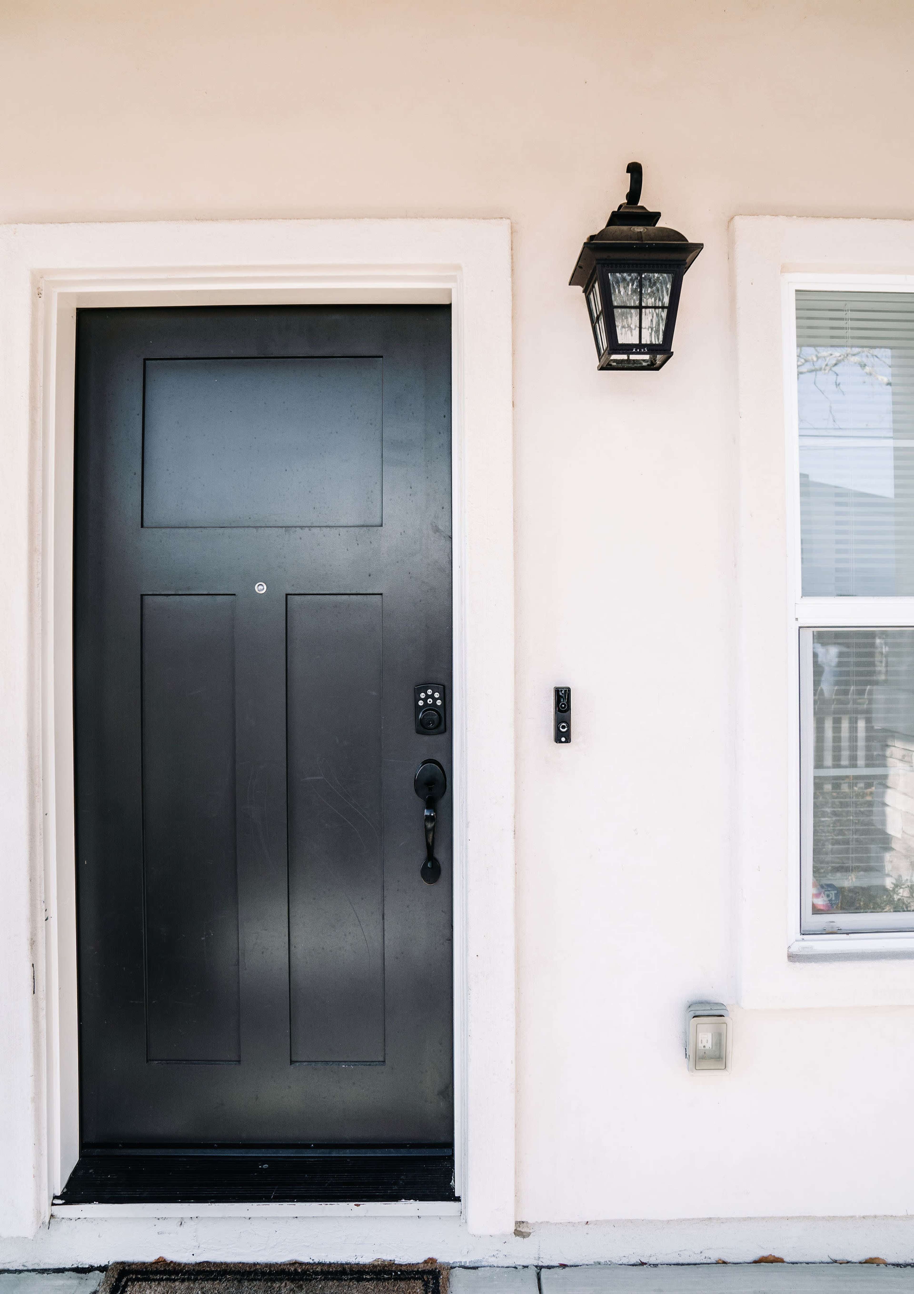 The entrance features a black door with a keypad lock and a lantern light attached to the side of a light-colored wall.