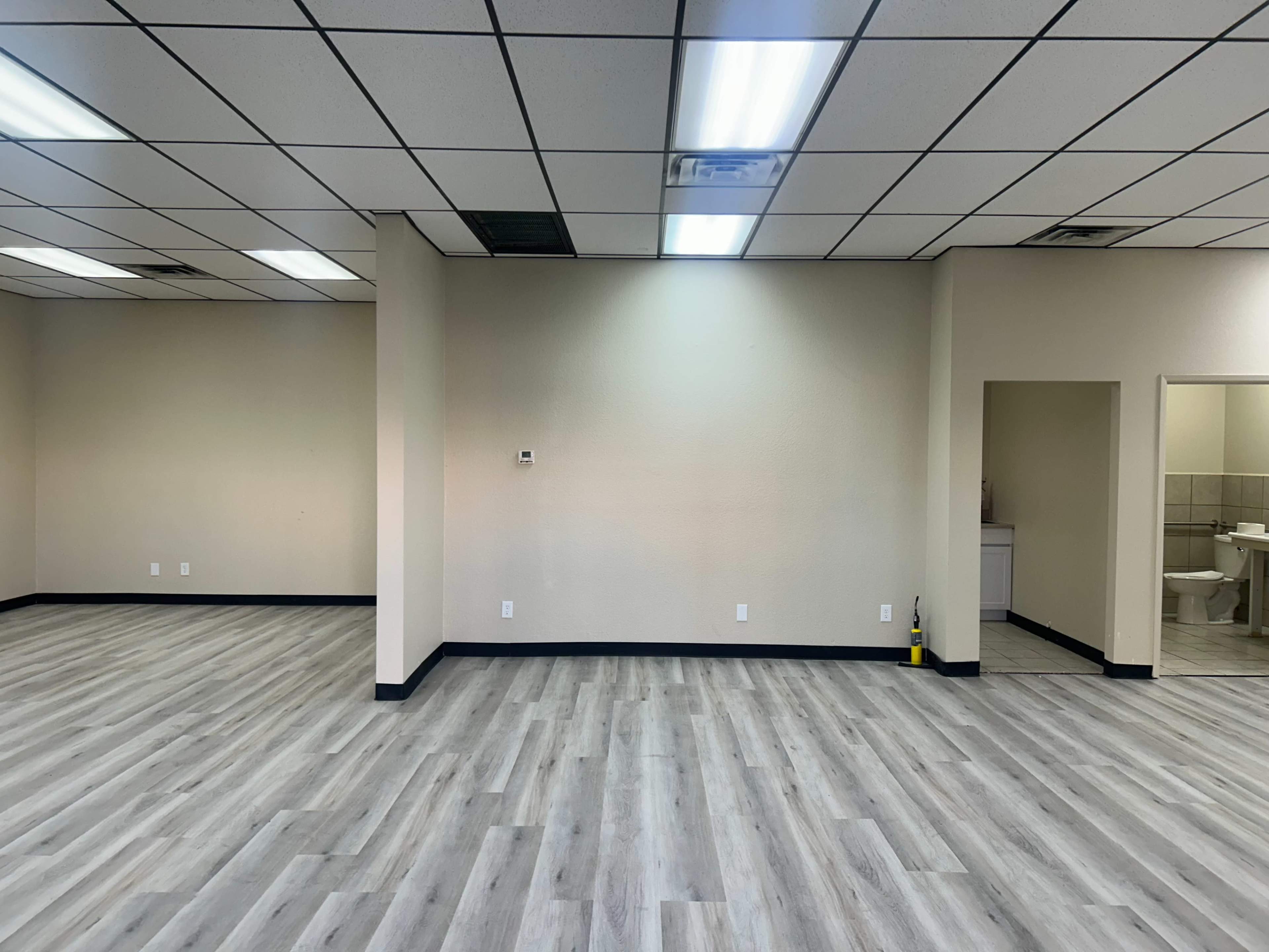 The image shows an empty commercial space with gray laminate flooring, plain walls, and a doorway leading to a bathroom area on the right.