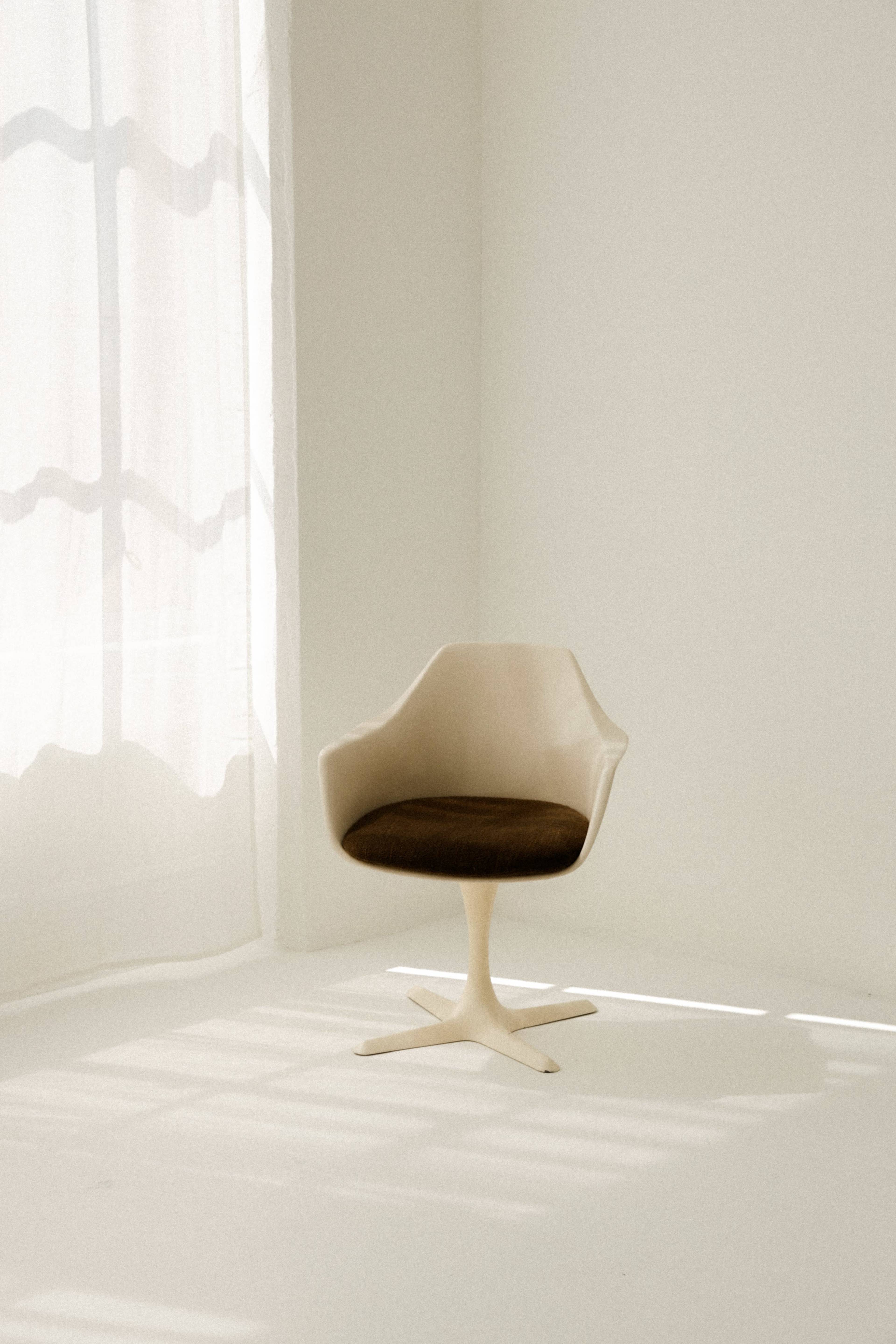 A white sculptural chair with a brown seat is positioned on a light-colored floor beside sheer curtains.