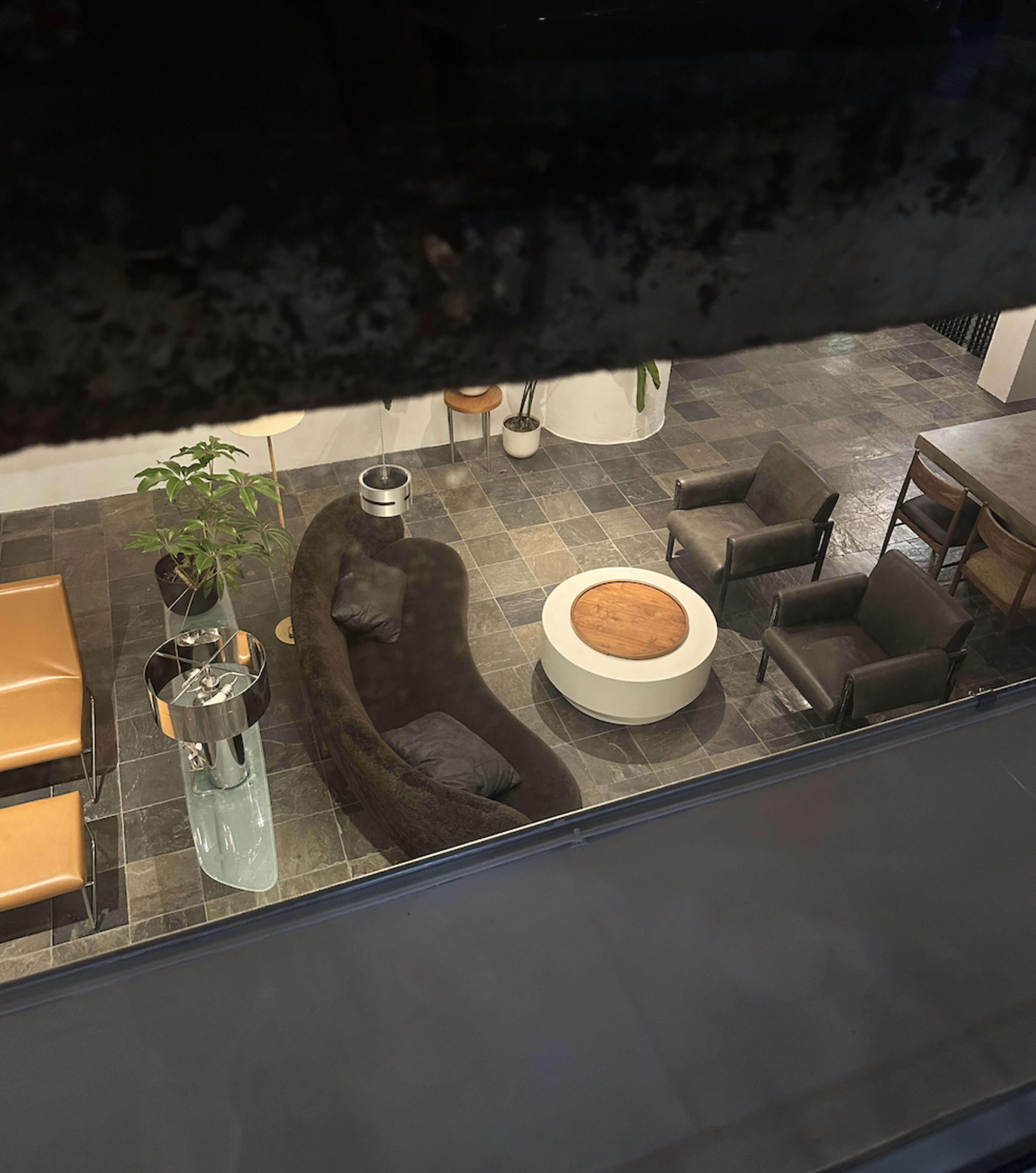 The image shows a modern lounge area featuring a brown sofa, several chairs, a round wooden coffee table, and a potted plant, all set on a tiled floor.