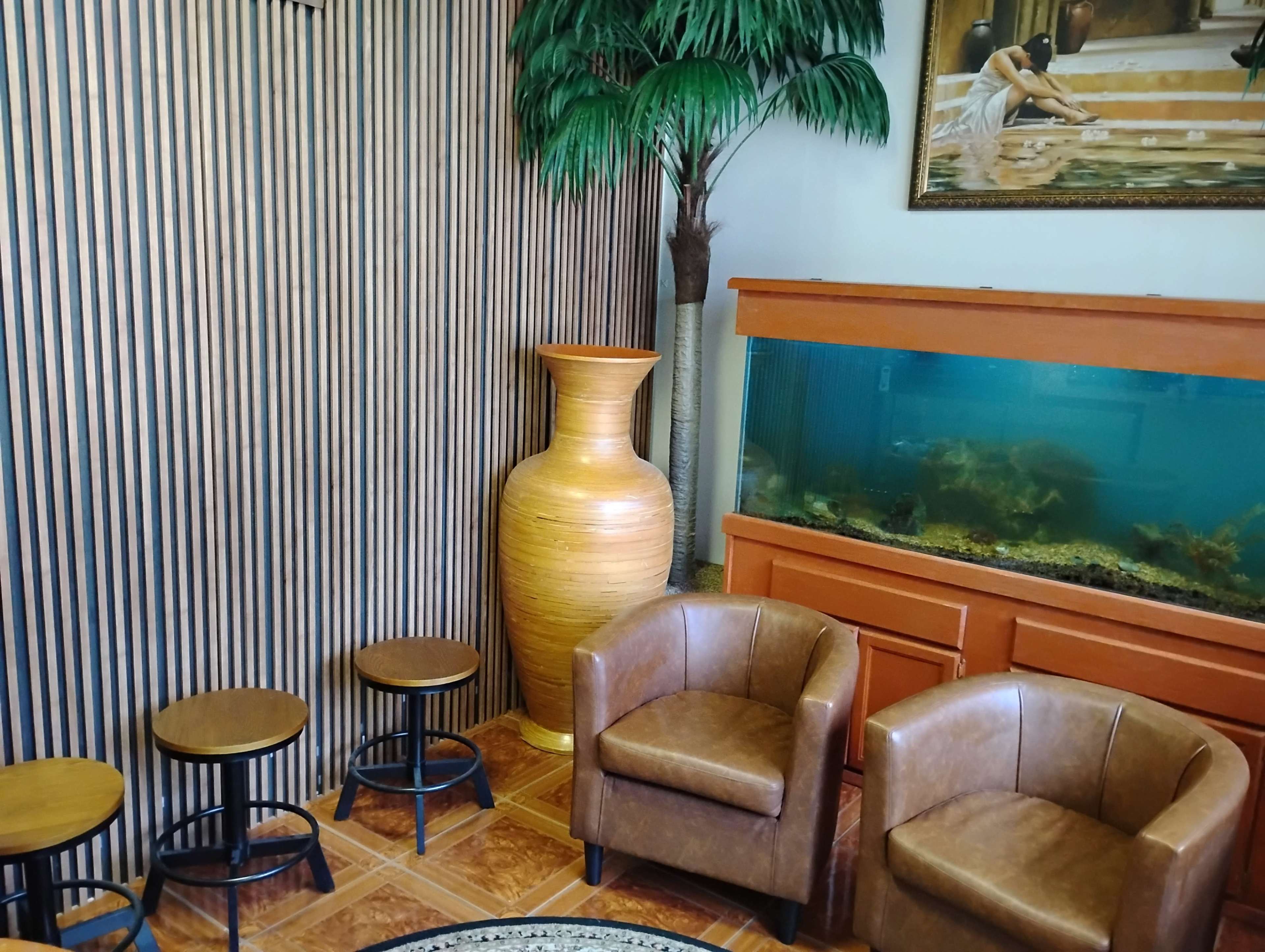 The image shows a small waiting area with two brown leather chairs, a large wooden vase, and an aquarium set against a backdrop of vertical wood paneling and a decorative plant.