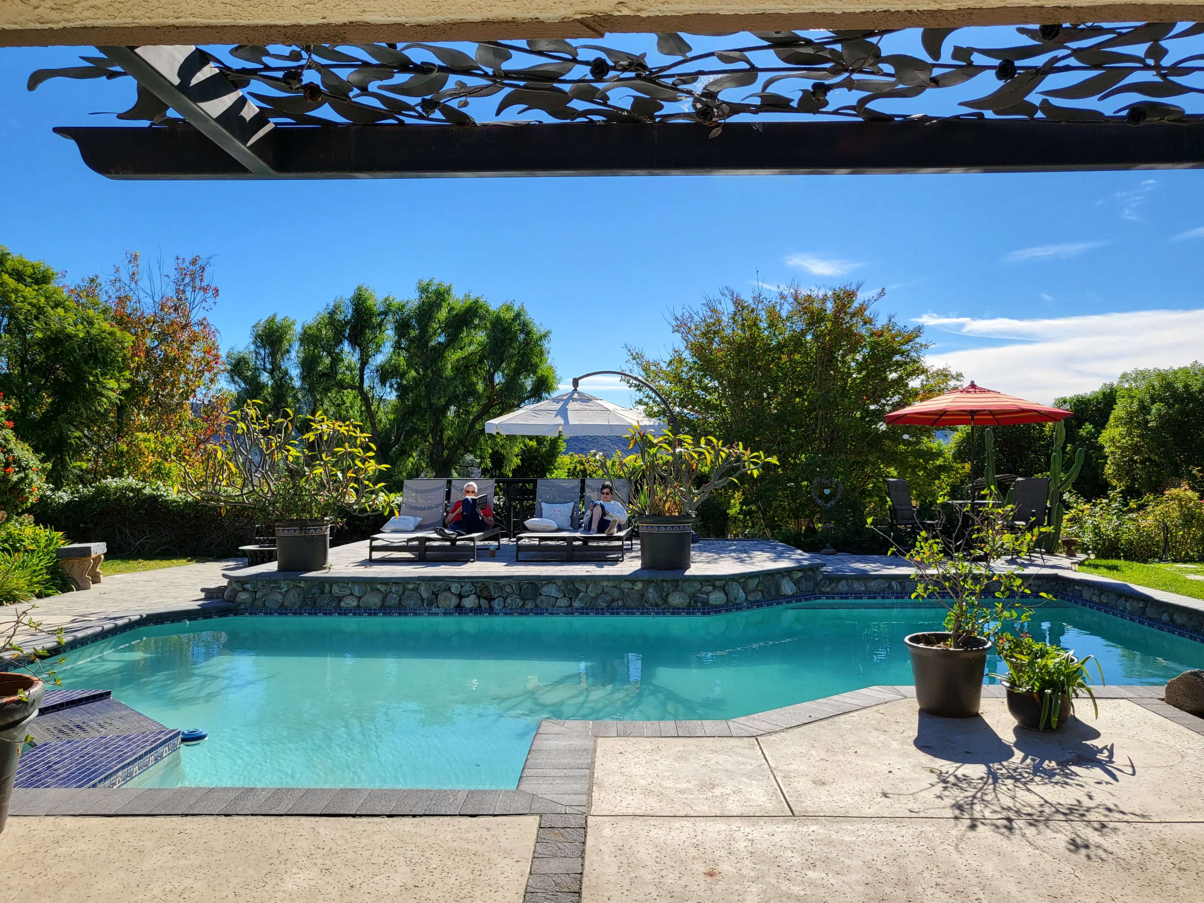 A pool area features lounge chairs, a sunshade, and surrounding greenery under a clear blue sky.