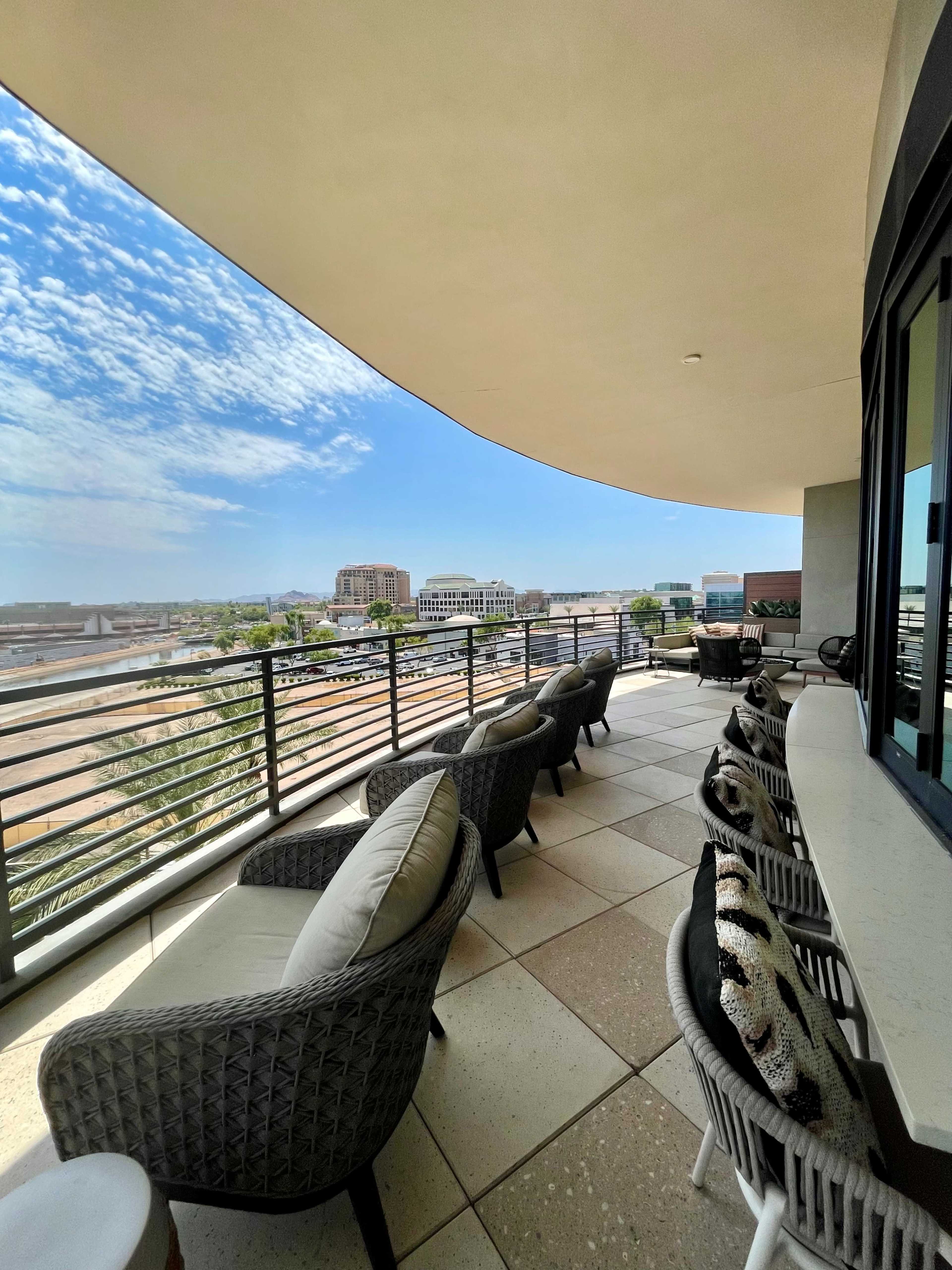The image shows a spacious balcony with modern seating and a view overlooking a cityscape under a clear blue sky.