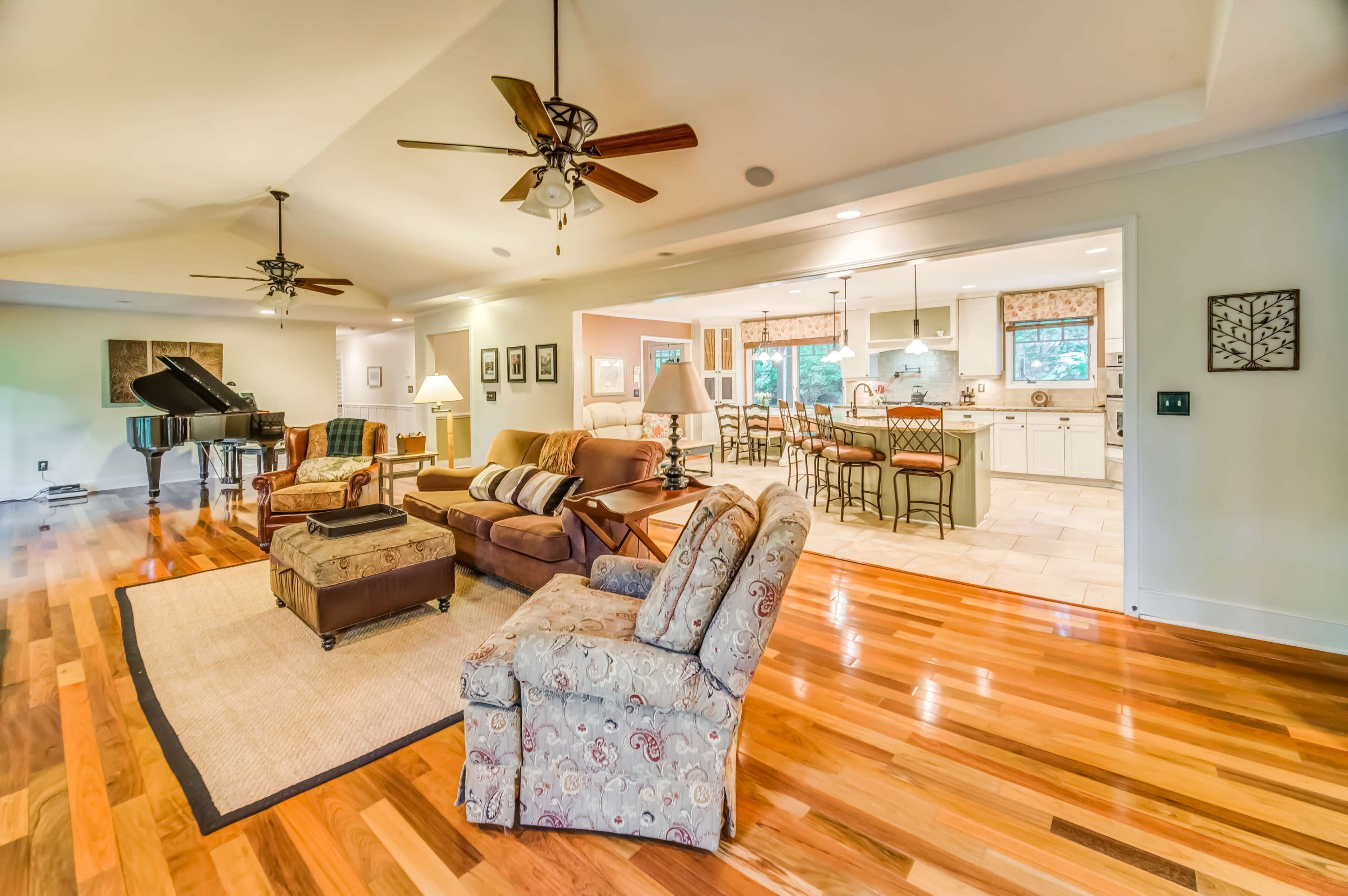 The image shows a spacious living area with hardwood floors, a piano, a seating arrangement with two armchairs, and a view of a kitchen area in the background.
