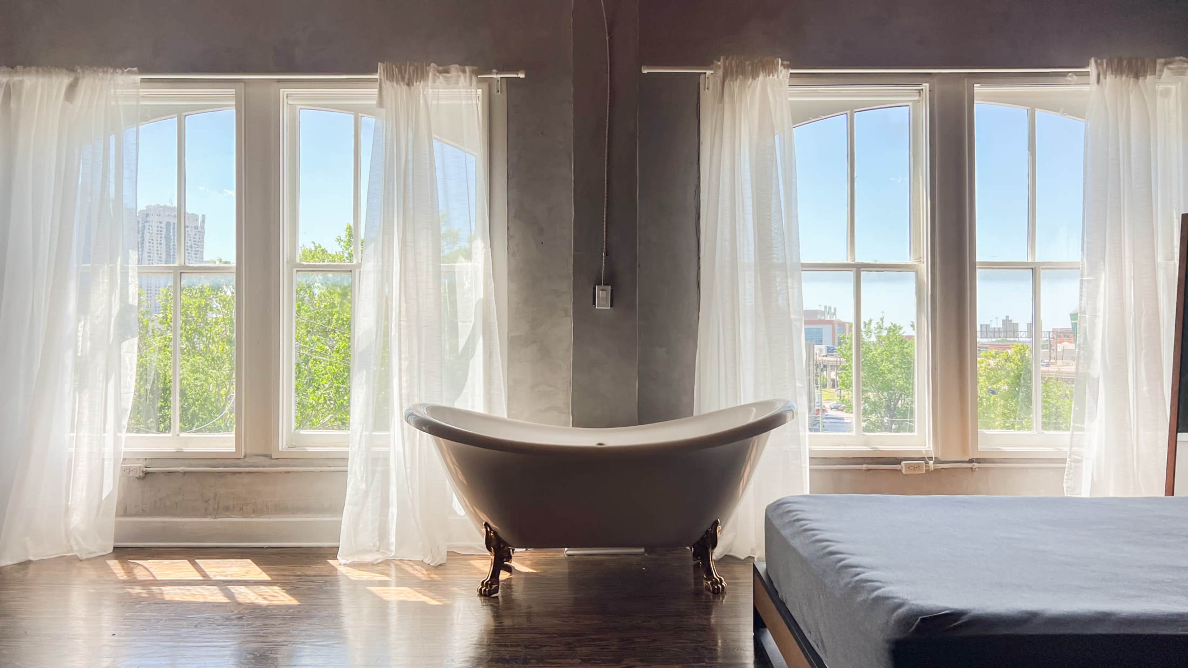 A freestanding white bathtub is positioned between two large windows with sheer curtains, allowing sunlight to illuminate the room with a view of greenery outside.