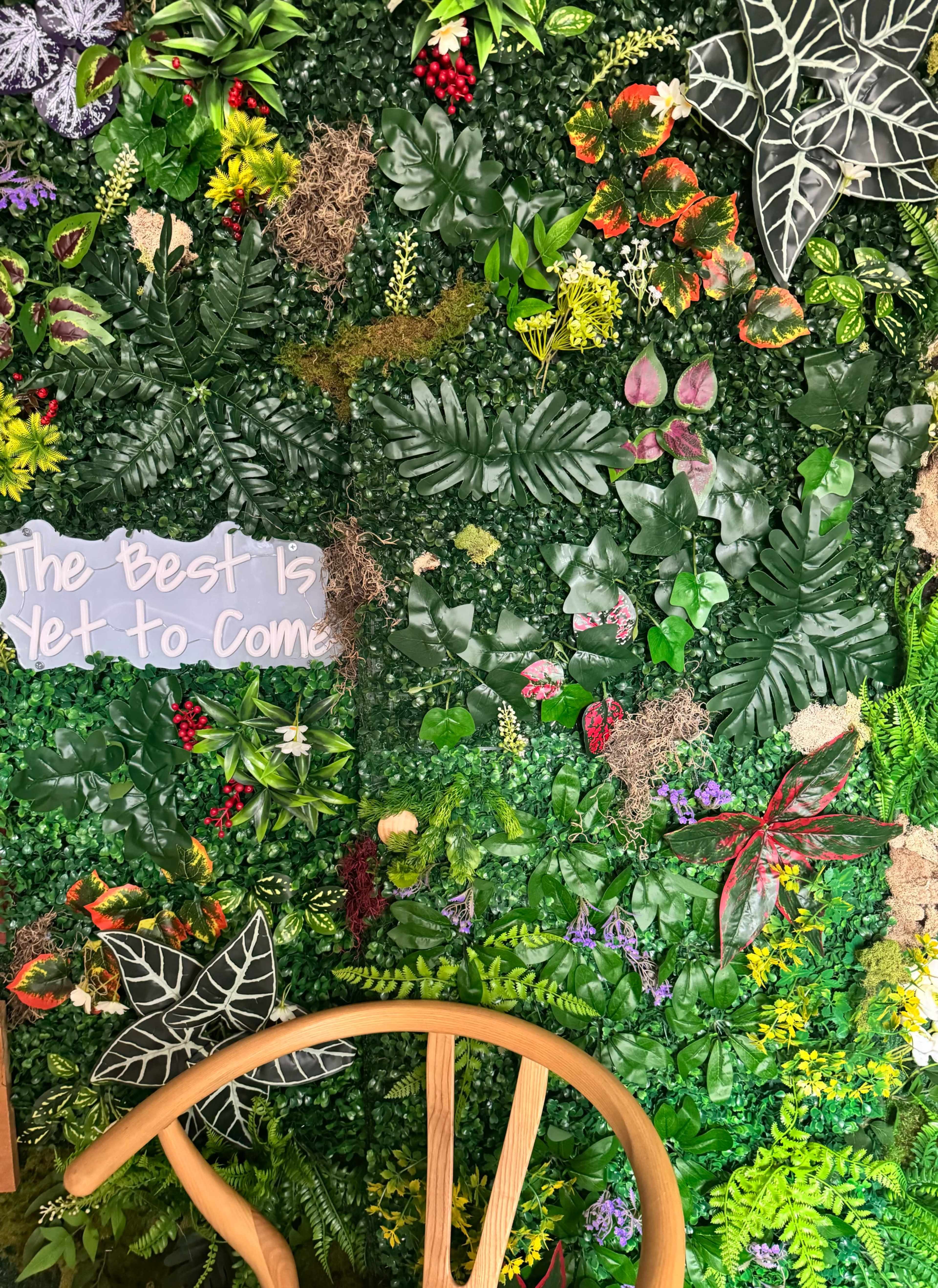 The image shows a vibrant wall covered in various artificial plants and leaves, along with a wooden chair in the foreground.