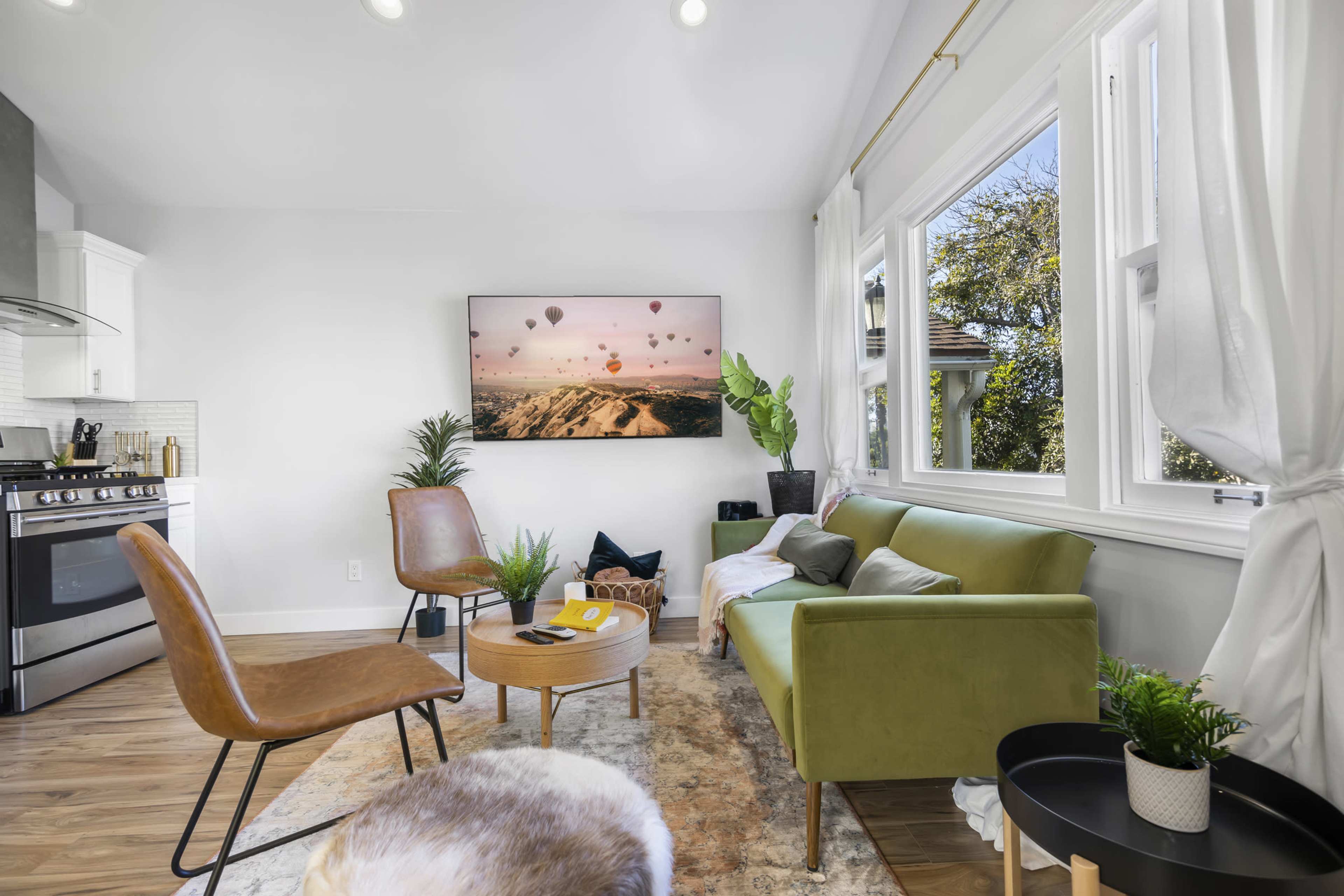 A modern living room features a green sofa, two chairs, a round coffee table, and a large wall-mounted television displaying a landscape with hot air balloons.